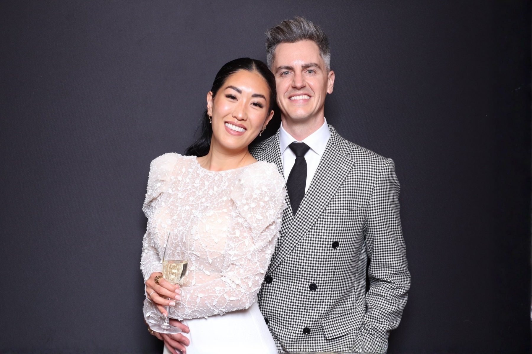 A smiling couple standing close together against a plain dark background, the woman holding a glass of champagne, both dressed in stylish formal attire.