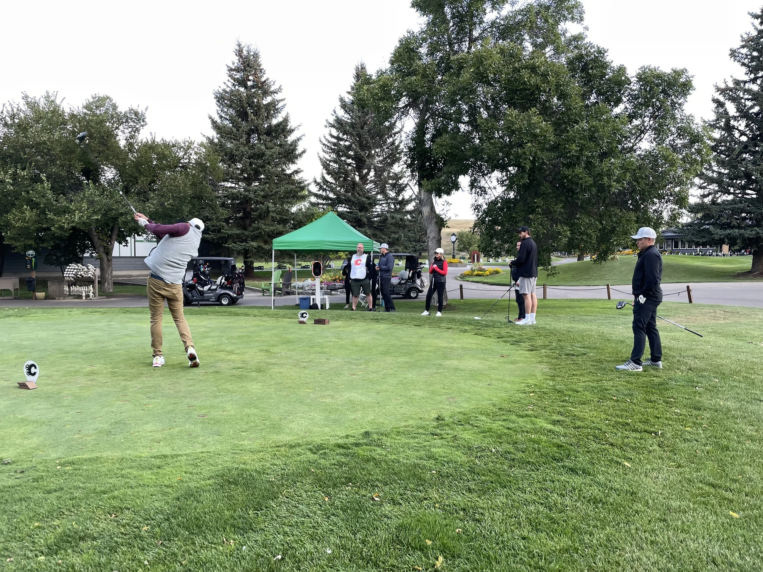 Group of people playing golf on a course with trees and a tent in the background.