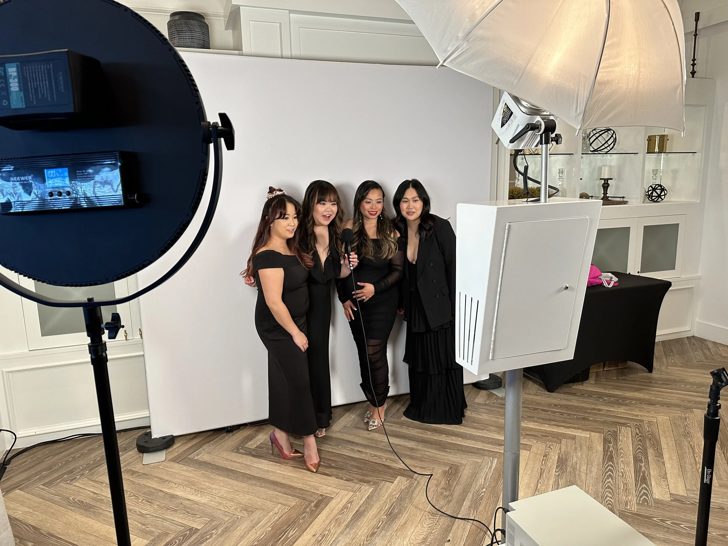 Four women dressed in black posing for a photo during a photo shoot in a room with white walls and wooden flooring. They are under a large umbrella light with photography equipment visible.