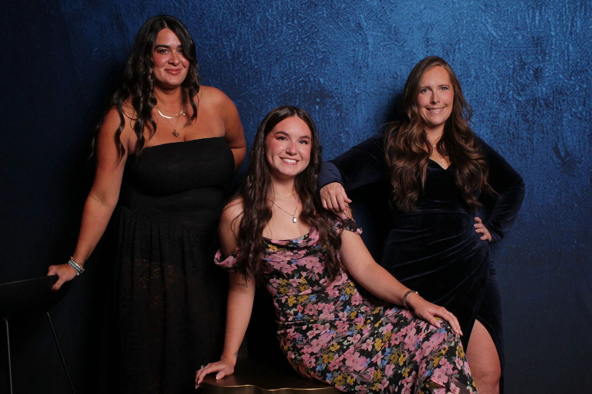 Three women posing in front of a dark blue textured wall, with one woman seated and two standing. They are dressed in elegant, dark-colored dresses and smiling at the camera.