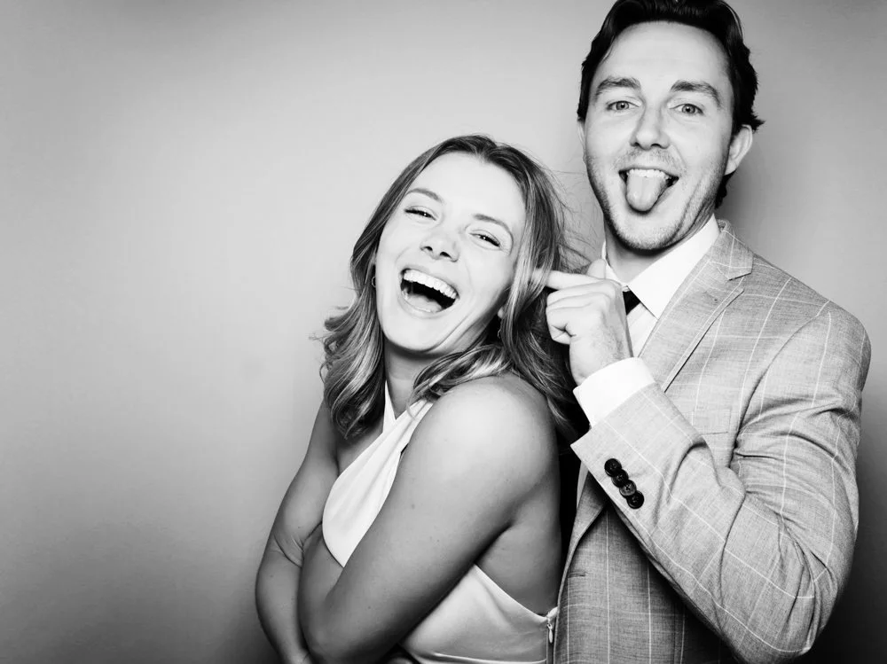 A man and woman in formal attire making silly faces and sticking out their tongues in a black-and-white photo