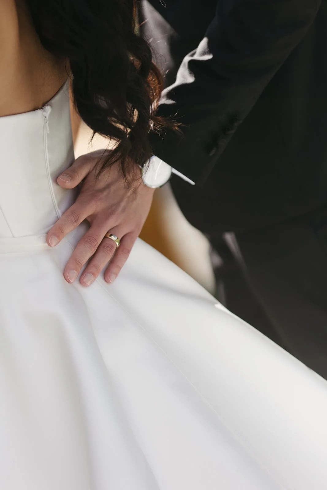 A person in a white dress, likely a wedding gown, is touching the hand of another individual who is dressed in a dark suit. The person in the white dress is wearing a ring on their left hand.