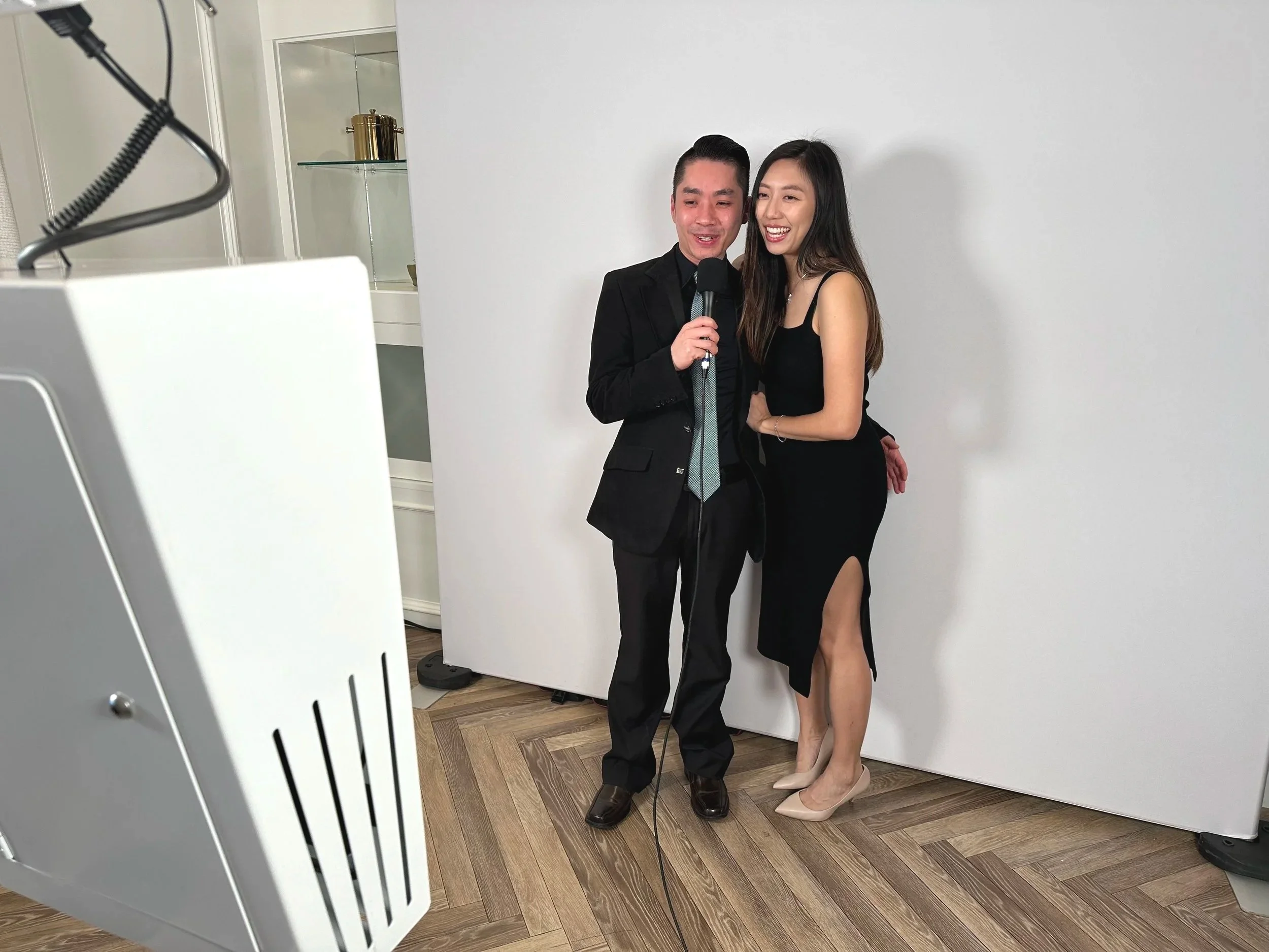 A man and woman are smiling and posing for a photo, with the man holding a microphone. They are dressed in formal attire, with the woman wearing a black dress and the man in a black suit. They are standing in front of a plain white backdrop, likely at an event or celebration.