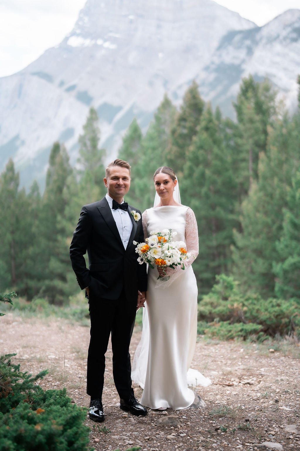 A bride and groom standing outdoors in a forested mountainous area, dressed in wedding attire, holding hands, with the bride holding a bouquet of white and orange flowers.