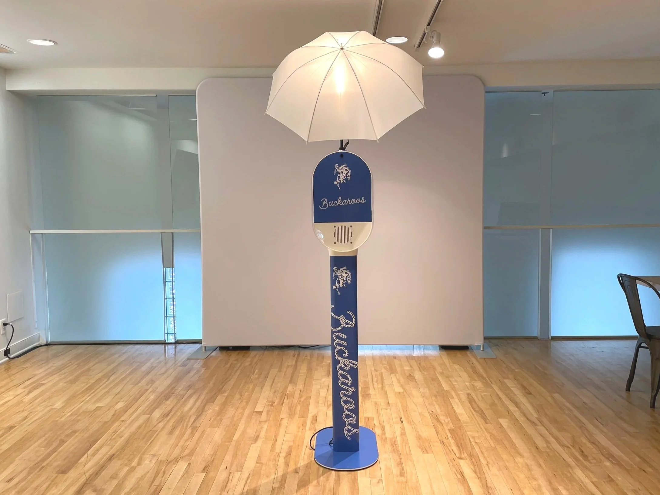 Lamp with a white umbrella shade on a blue stand featuring Buckaroo logo and text, placed in a modern indoor space with hardwood flooring and frosted glass walls.