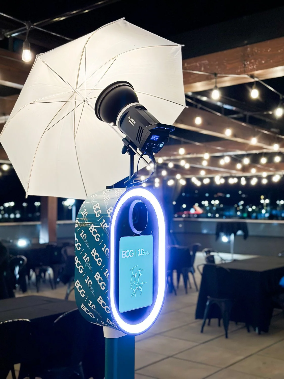 A digital check-in station with a white illuminated ring, a screen prompting 'Tap to Start', an attached USB charger, a white umbrella, a camera mounted above, and a venue with outdoor seating at night in the background.