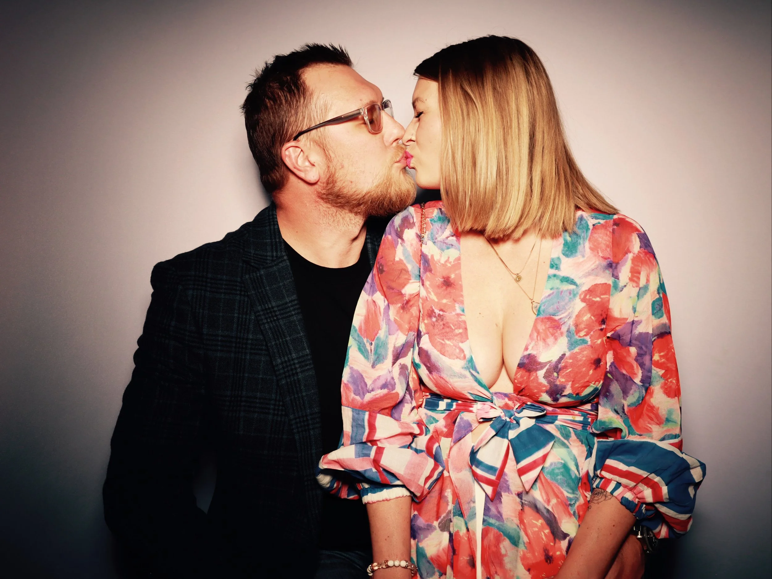 A man and woman are about to kiss, facing each other with noses touching. The man has short dark hair, glasses, facial hair, and is wearing a dark jacket. The woman has shoulder-length blond hair and is wearing a colorful, floral dress with a deep ne