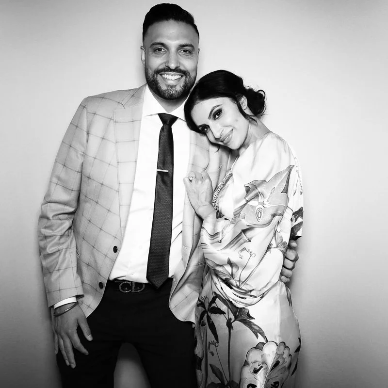 A man and woman standing close together, smiling, in front of a plain background. The man is wearing a light-colored checked blazer, white shirt, and dark tie. The woman is wearing a floral-patterned dress and has her head resting on the man's should
