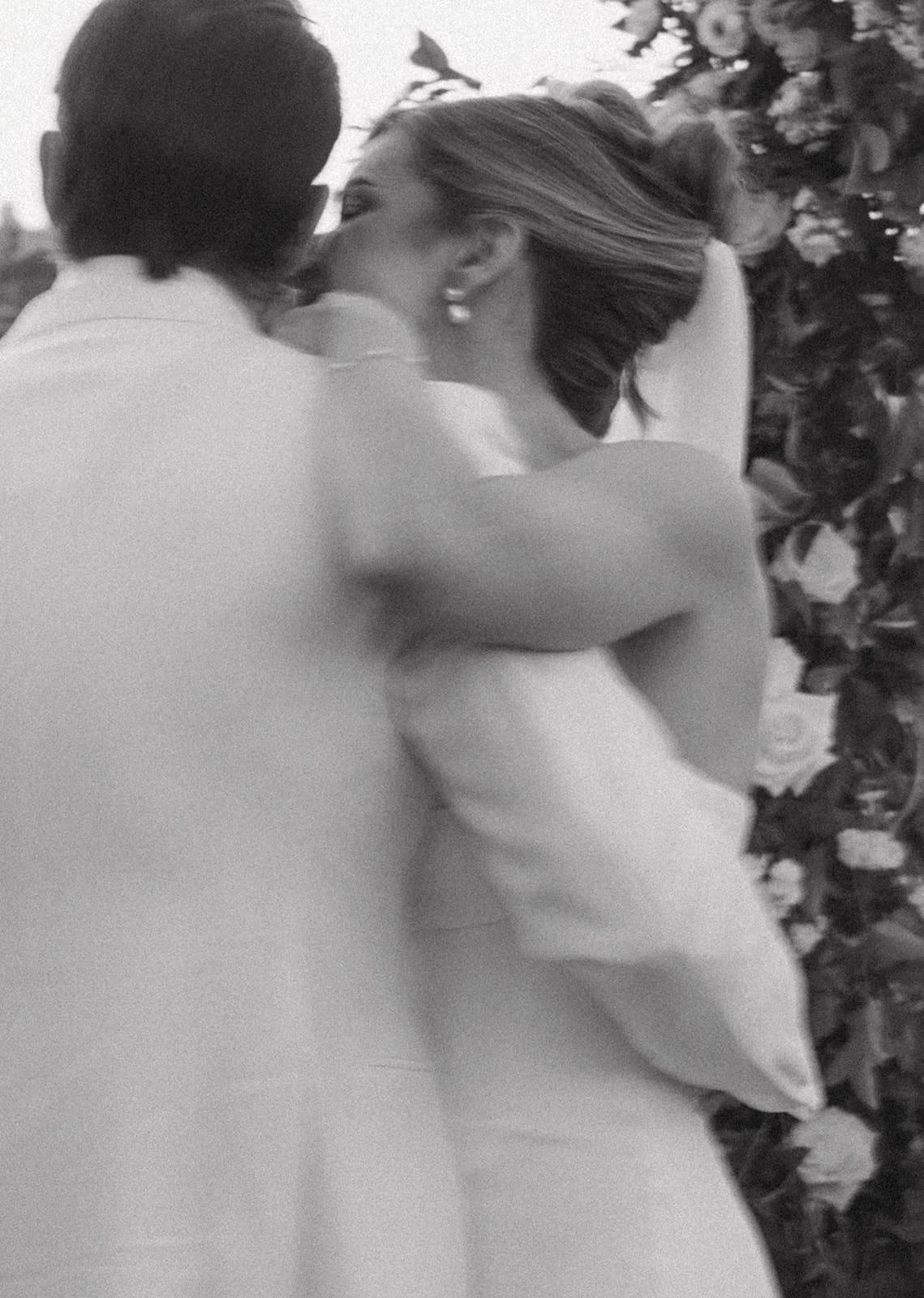 A black and white photo of a couple sharing a kiss, with the man facing away and the woman facing sideways, surrounded by flowers.