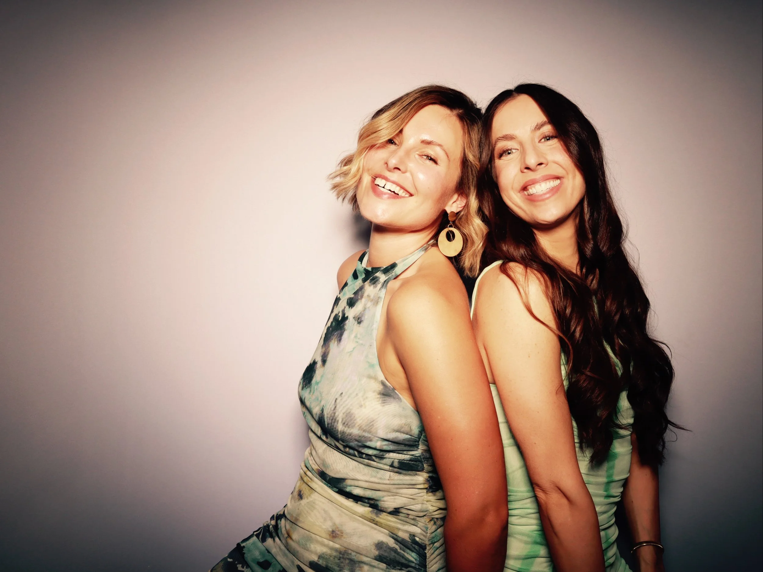 Two women smiling and standing back to back, wearing colorful dresses, against a plain background.