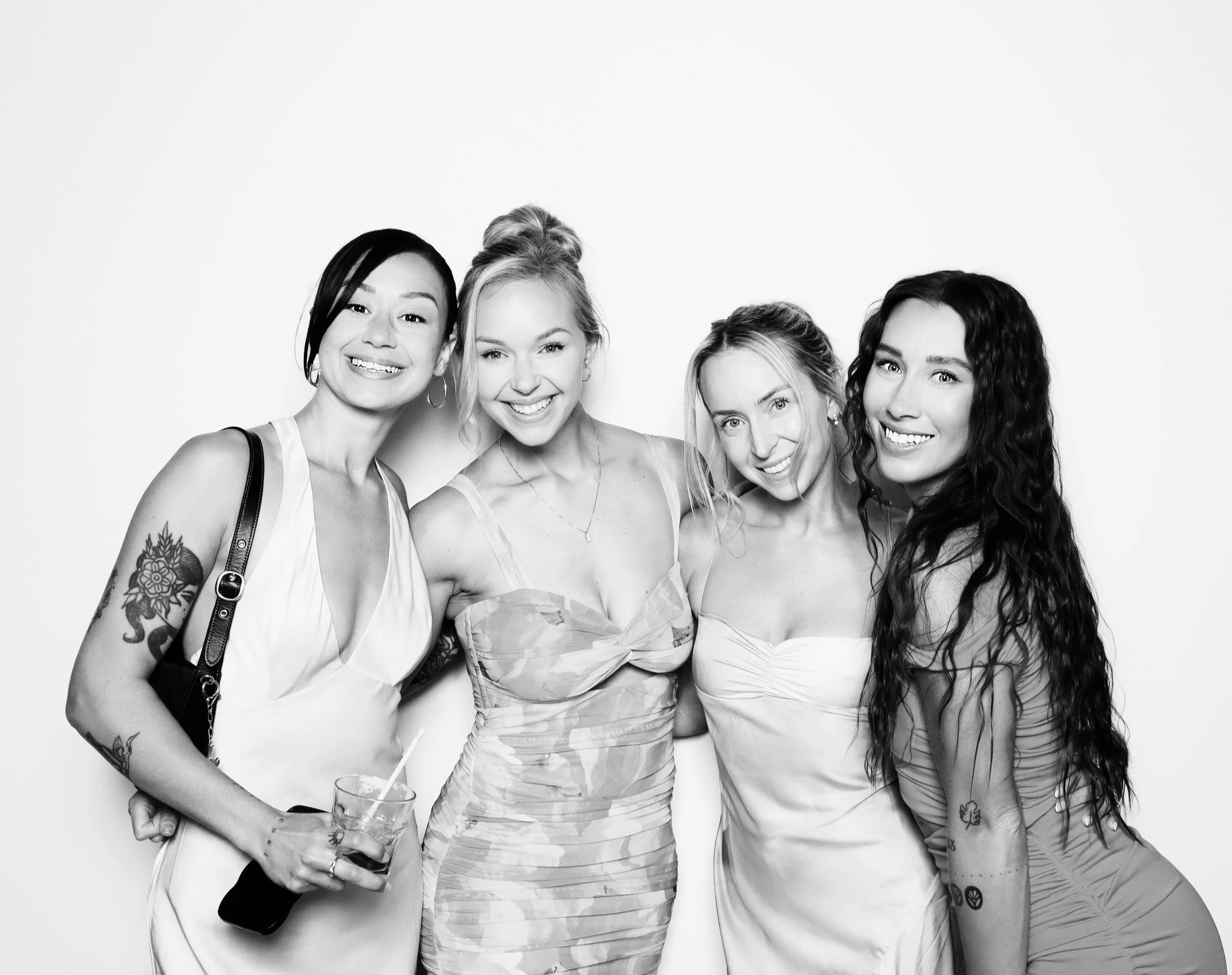 Four women standing close together, smiling, posing for a photo in front of a plain background. One woman has tattoos and is holding a drink and a phone. They are all dressed in summer or party attire.