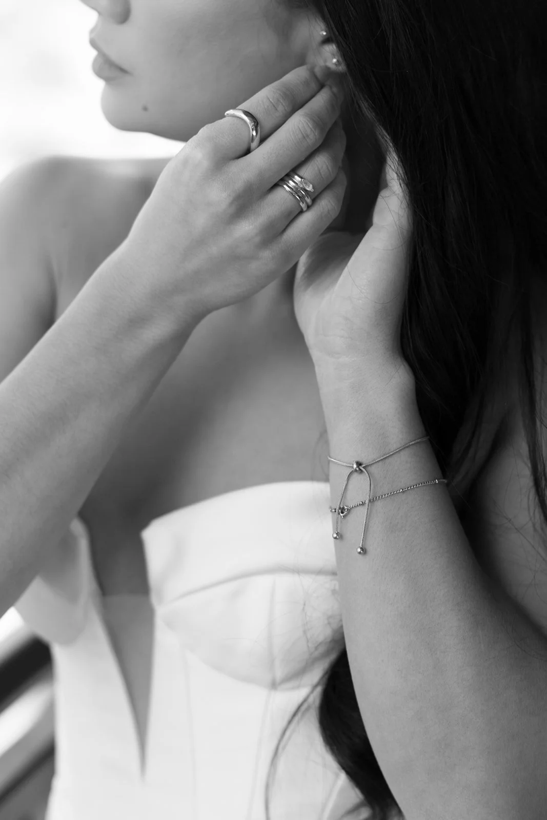 Black and white close-up of a woman putting on earrings, wearing multiple rings and bracelets.