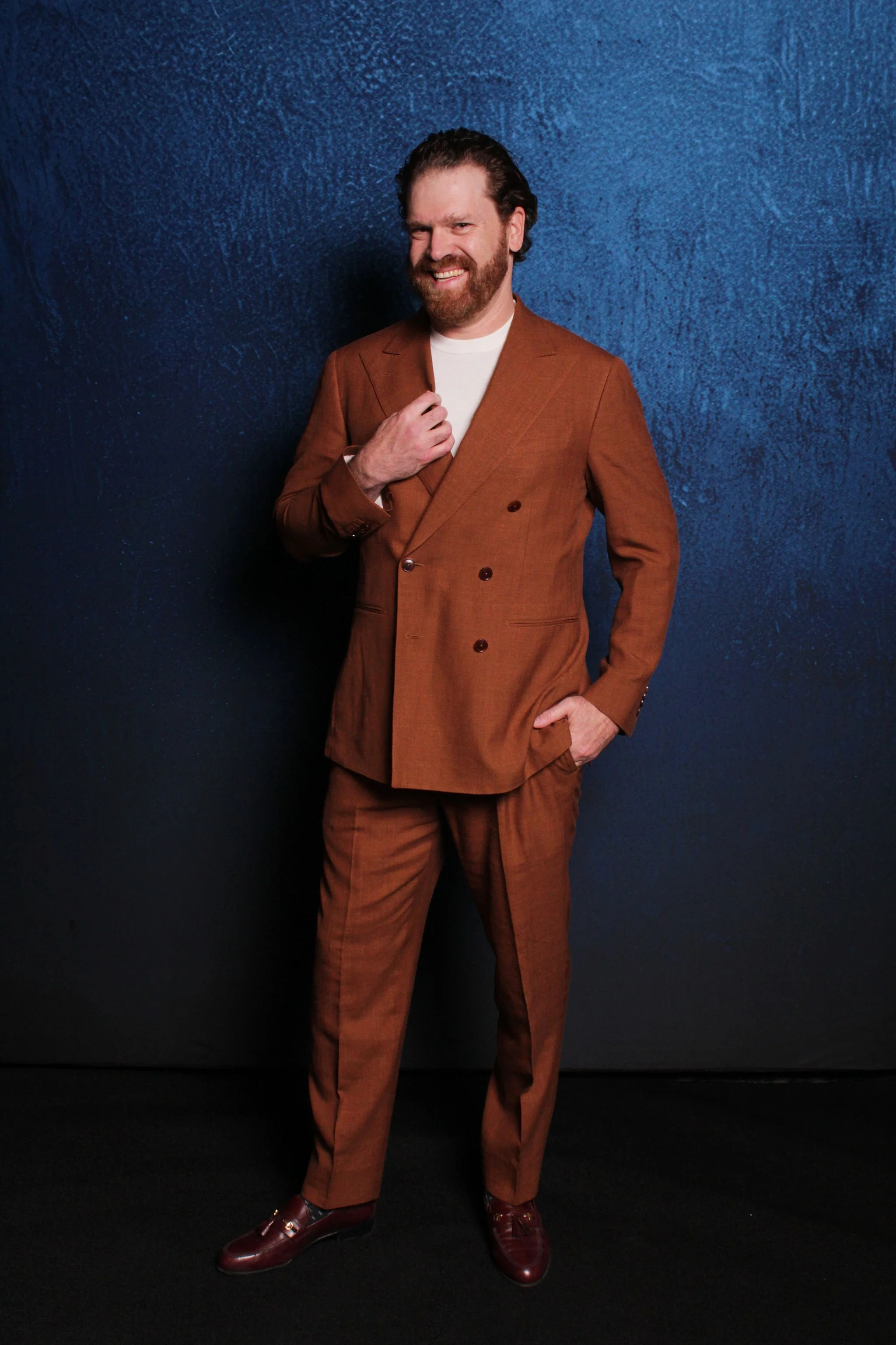 A man with a beard and styled hair, wearing a brown double-breasted suit and brown shoes, standing against a dark blue textured wall. He is smiling, with one hand touching his suit lapel and the other in his pocket.