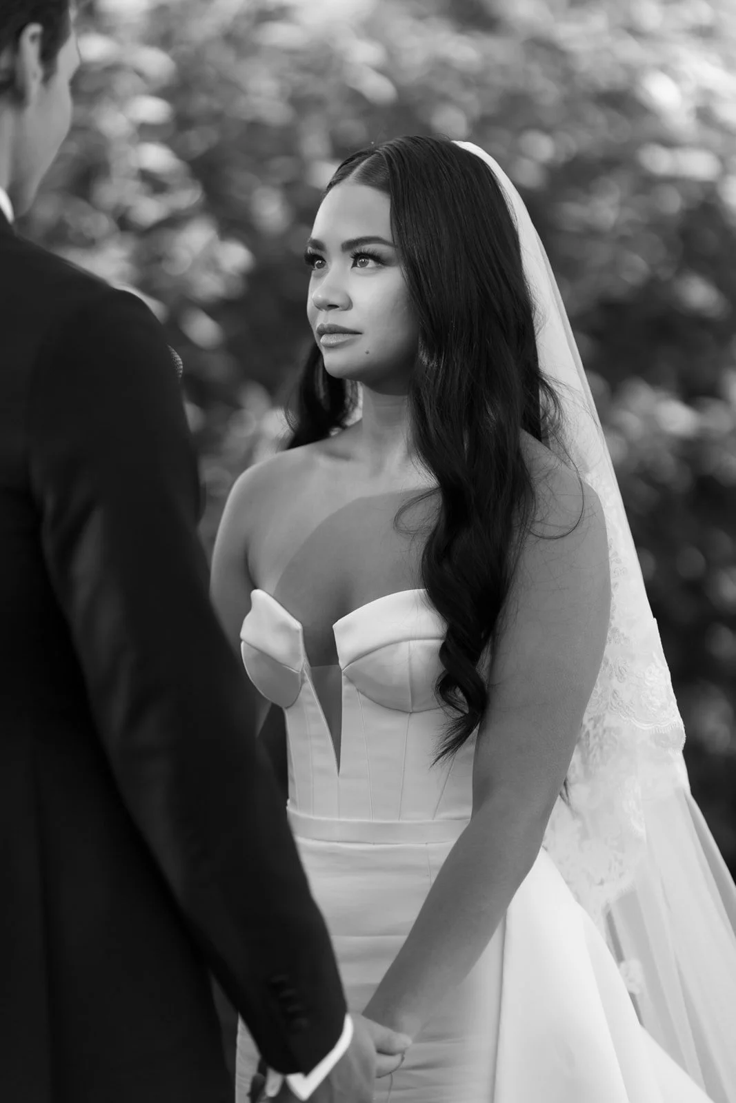 A bride with long dark hair and a strapless wedding dress looking at her groom during their wedding ceremony outdoors.