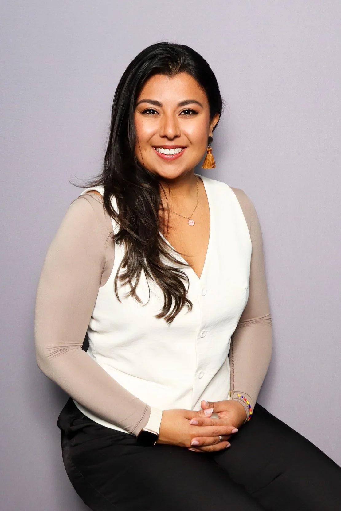 A woman with long dark hair, wearing a beige and white blouse, sitting against a plain gray background, smiling at the camera.