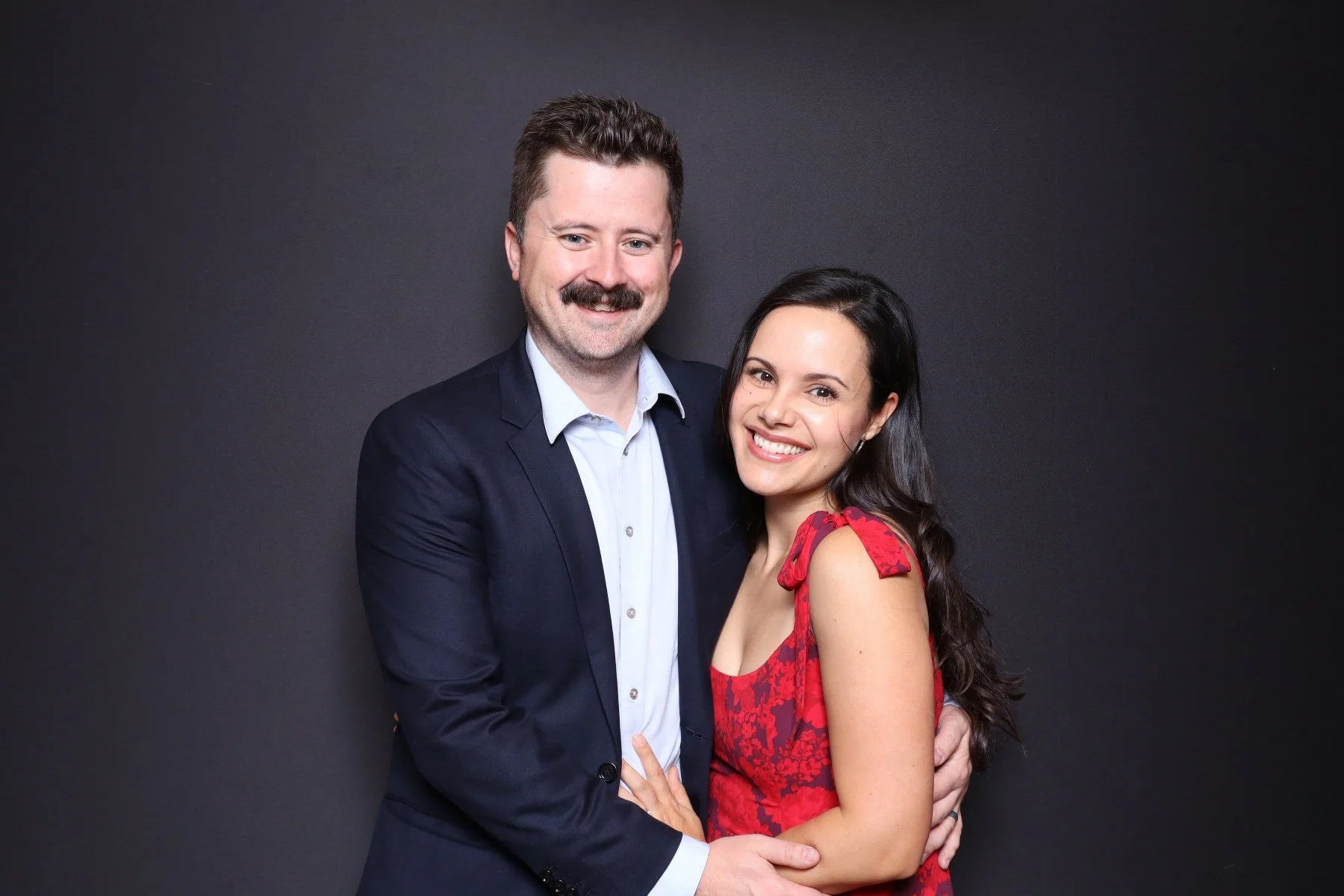 A man in a dark suit with a light blue shirt and a woman in a red dress with shoulder ties pose happily together against a black background.