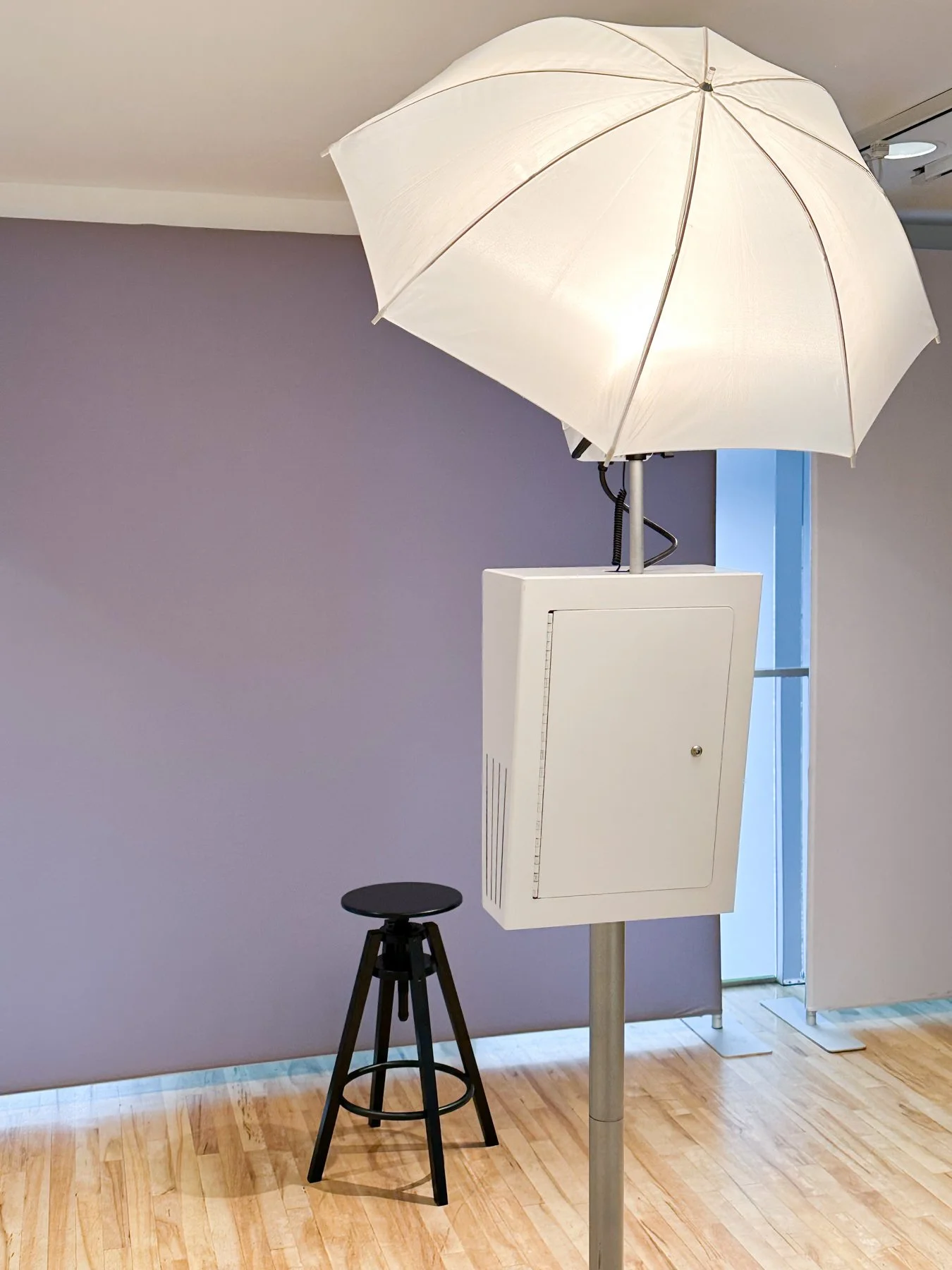 A photography setup with a white umbrella light, a black stool, and a white electrical box on a stand inside a room with a wooden floor and a purple wall.