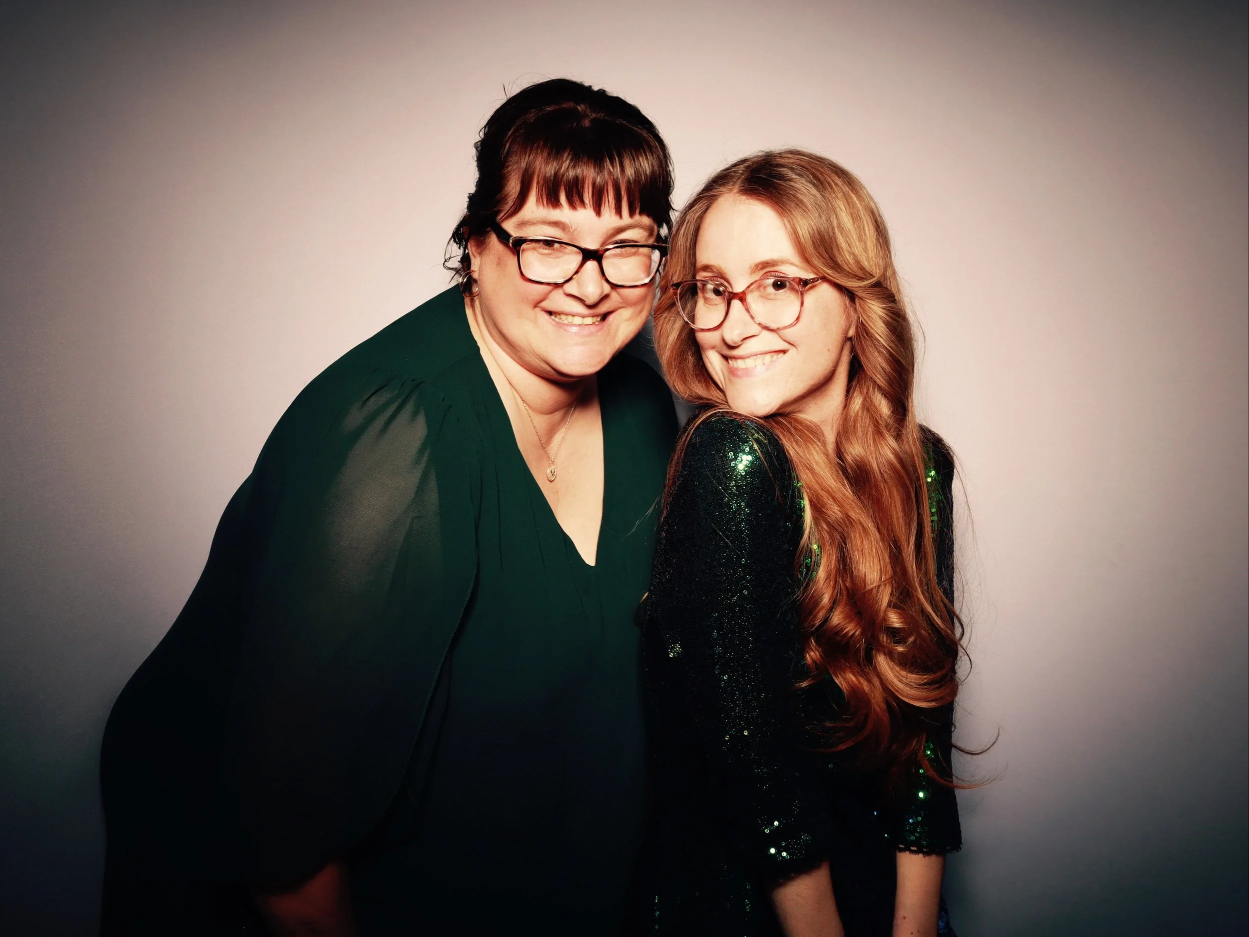 Two women stand close together, smiling at the camera. The woman on the left has short dark hair, glasses, and is wearing a dark green top. The woman on the right has long red hair, glasses, and is wearing a sparkly black top.
