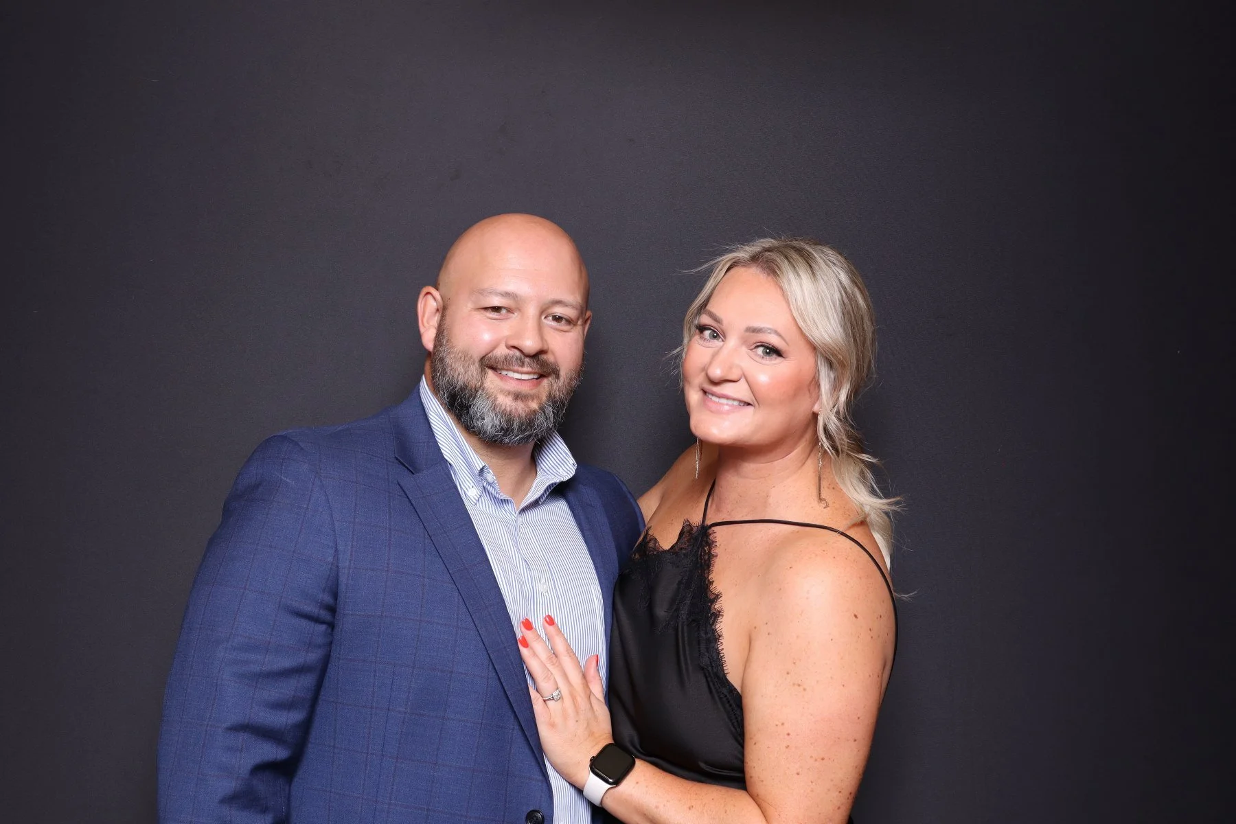 A smiling couple standing close together against a dark background. The man has a beard, is bald, and is wearing a blue checked suit with a white dress shirt. The woman has blonde hair, is wearing a black sleeveless dress with thin straps, and has he