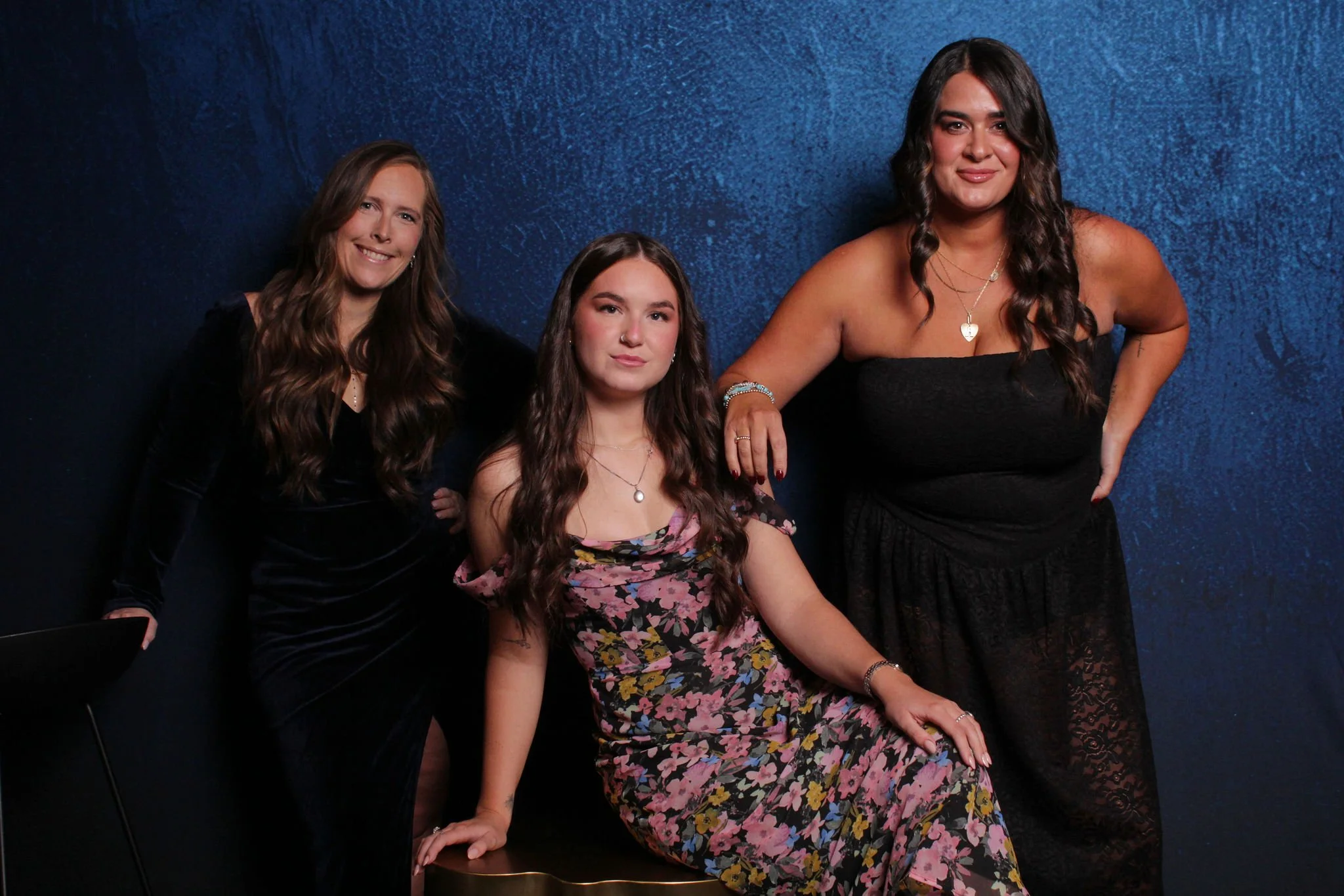 Three women posing against a dark blue textured background. The woman on the left has long wavy hair and is wearing a black dress. The woman in the center has long wavy hair and is sitting on a chair, wearing a floral dress. The woman on the right ha