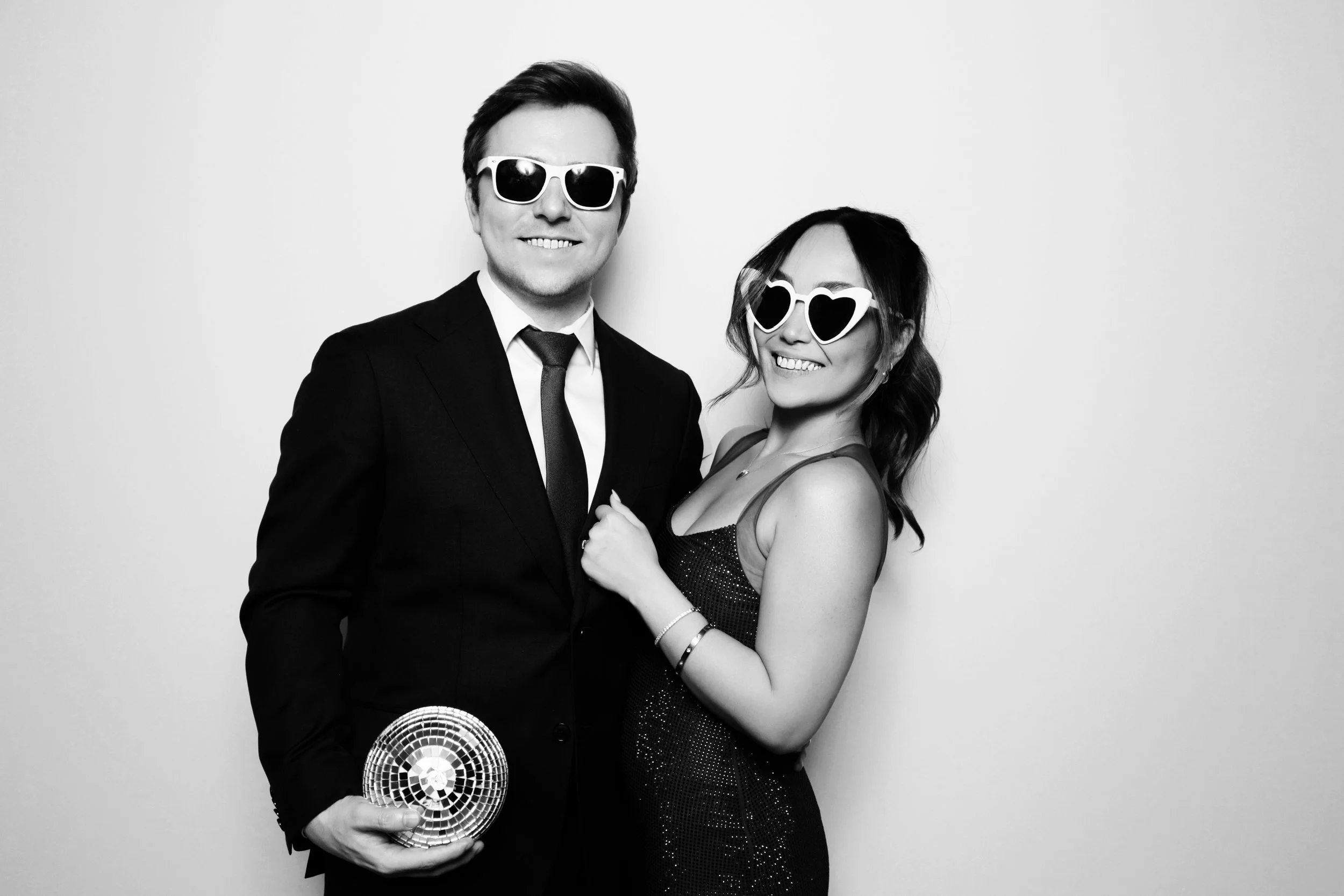 A man and woman in formal attire wearing sunglasses and holding a disco ball, smiling against a plain background.