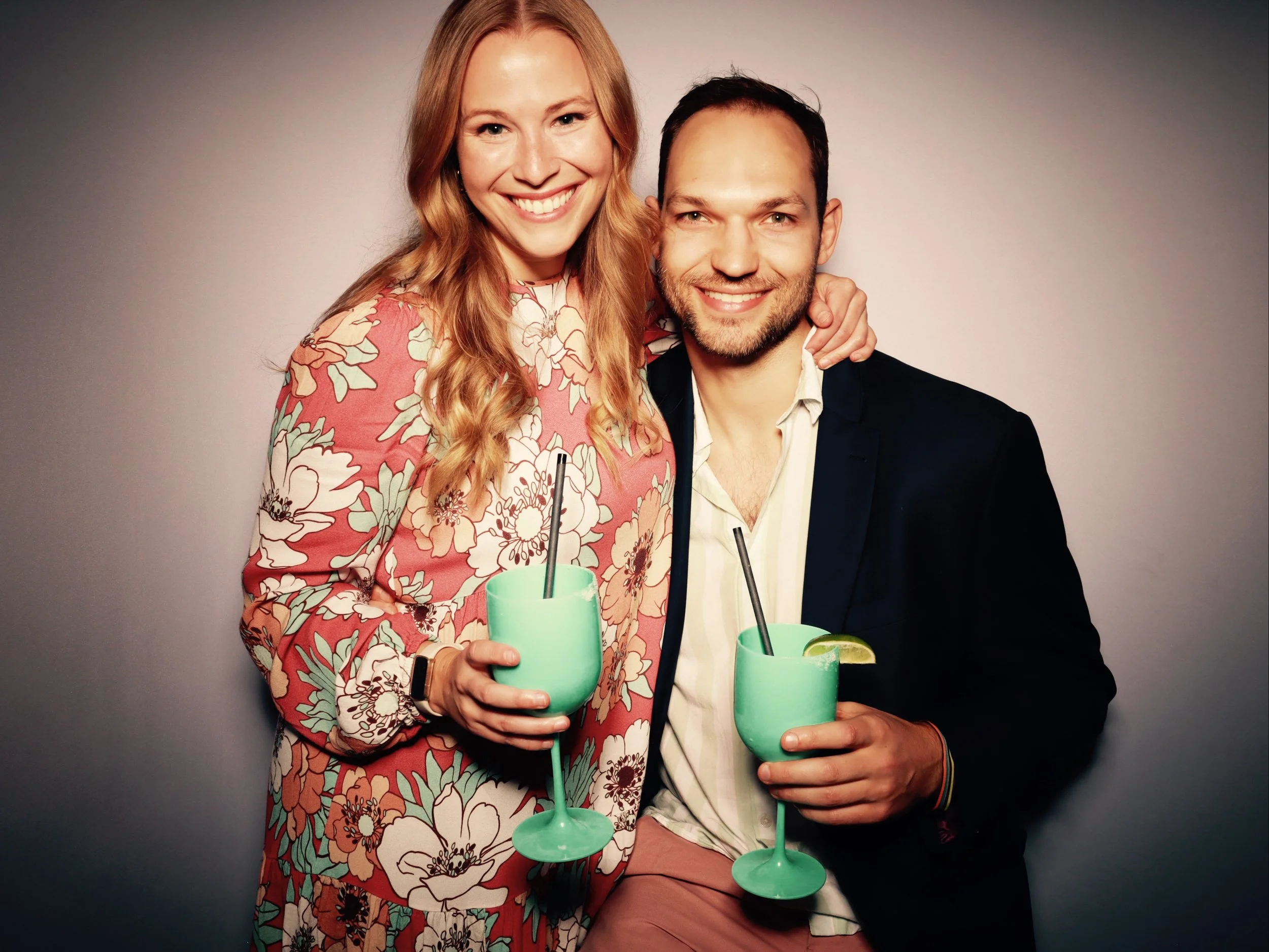 A smiling woman with long red hair wearing a floral patterned dress holding a blue cocktail, standing next to a smiling man with dark hair and beard wearing a dark blazer and a light-colored shirt, holding a blue cocktail with a lime wedge, both in f