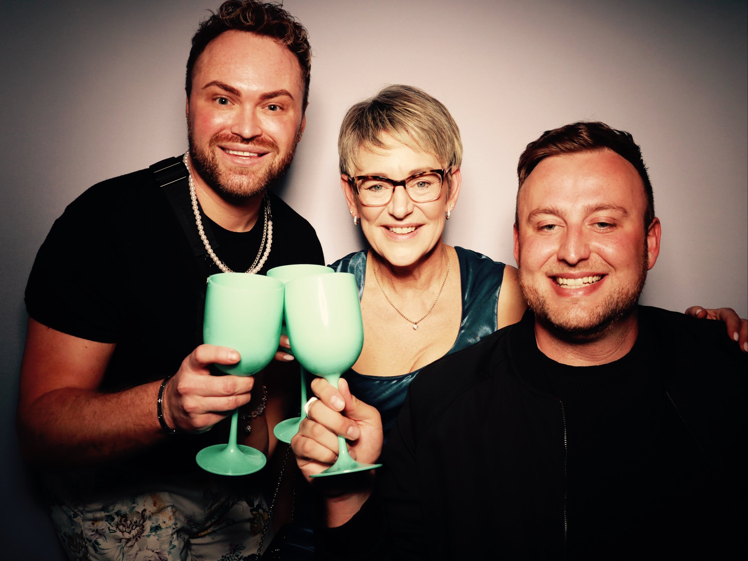 Three people, two men and one woman, smiling and holding green glasses, celebrating together in front of a plain gray background.