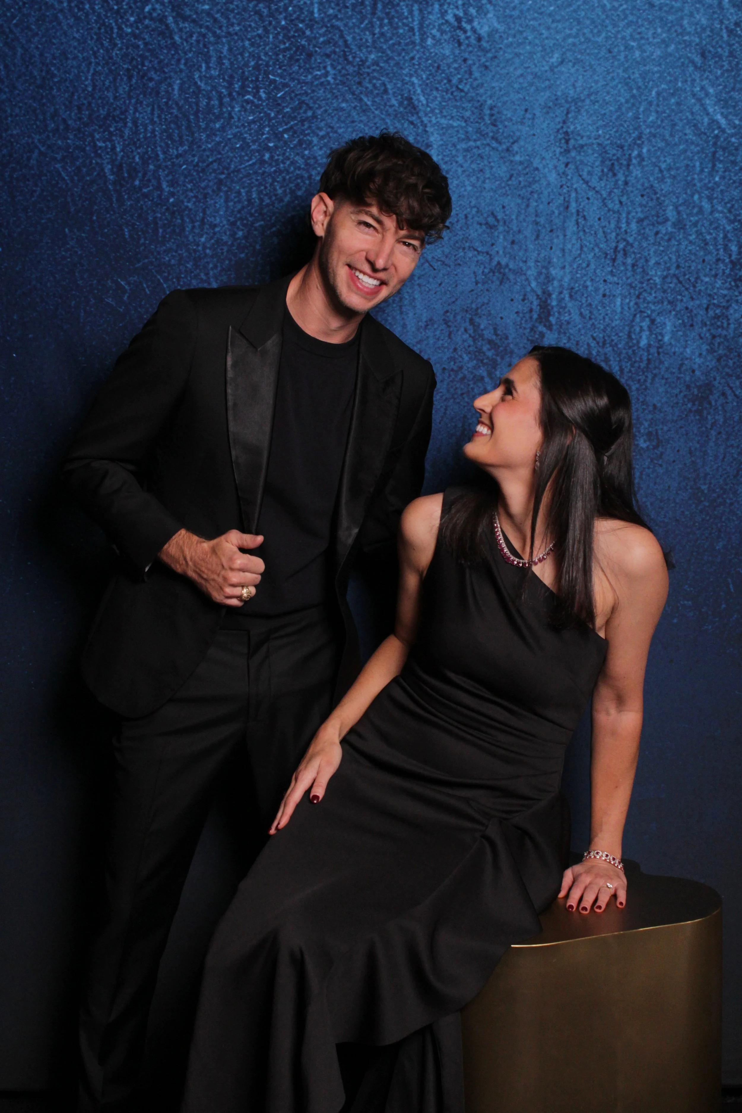 A man in a black tuxedo with a satin lapel and a woman in a black evening gown sharing a joyful moment against a textured dark blue background.