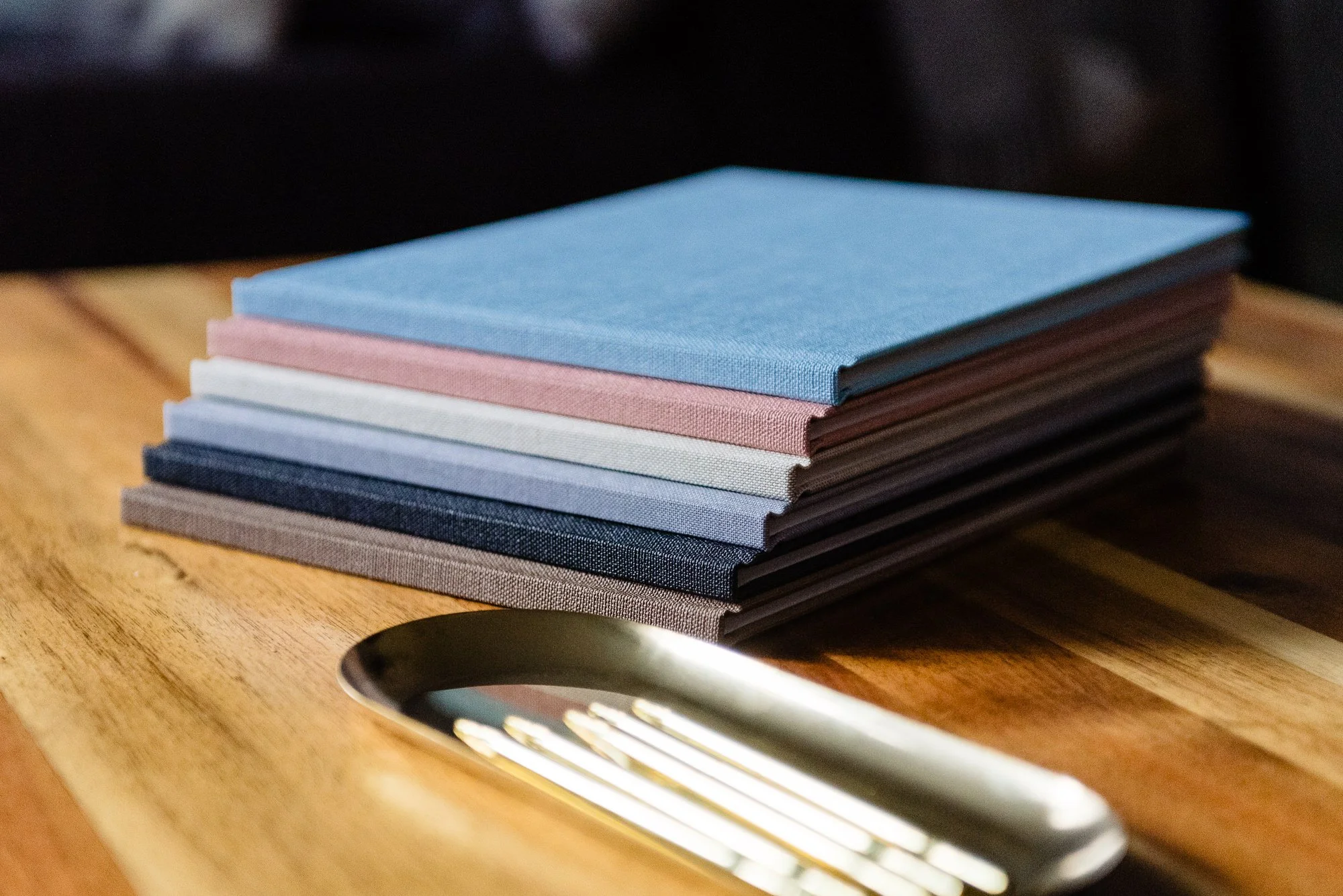 Stack of several colorful books on a wooden table beside a silver utensil, possibly a spoon.
