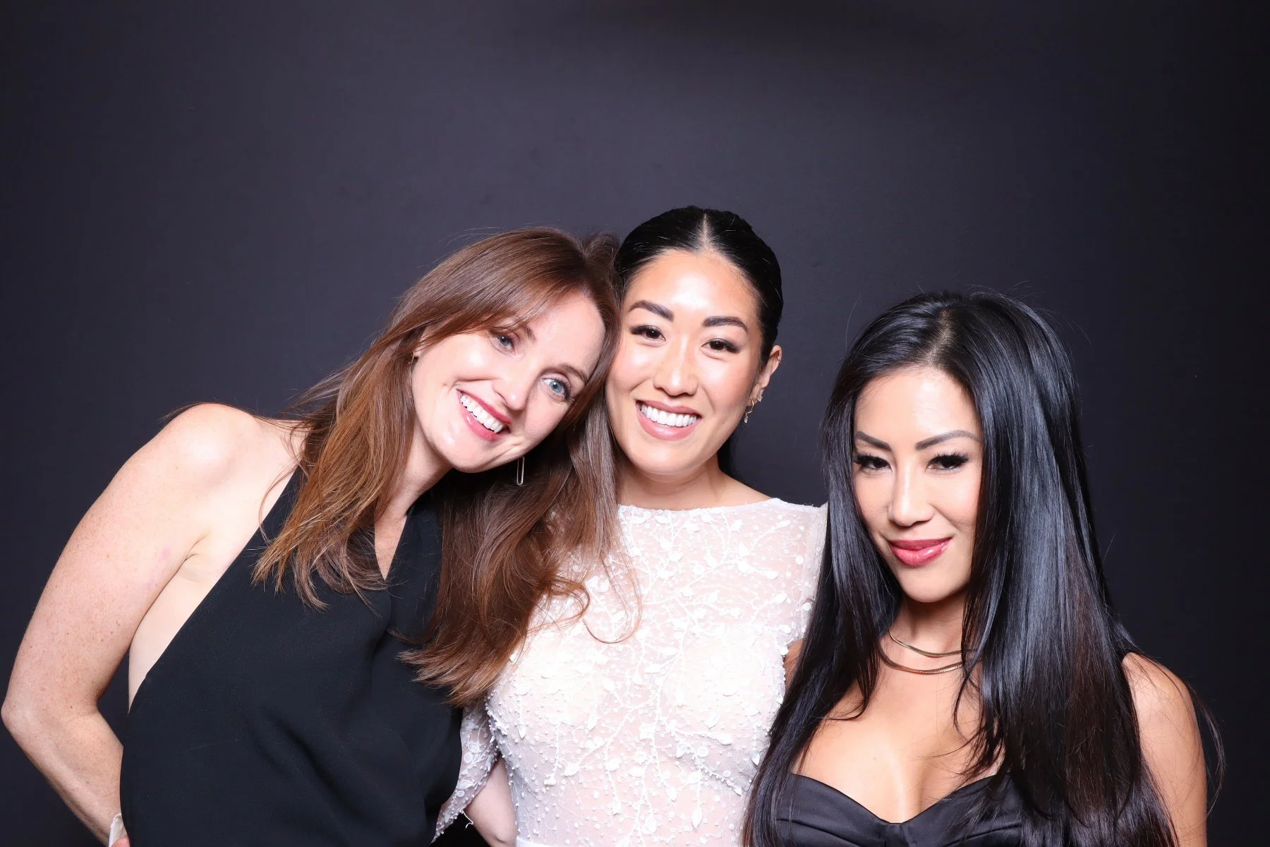 Three women smiling and posing against a black background, dressed elegantly for a formal occasion.