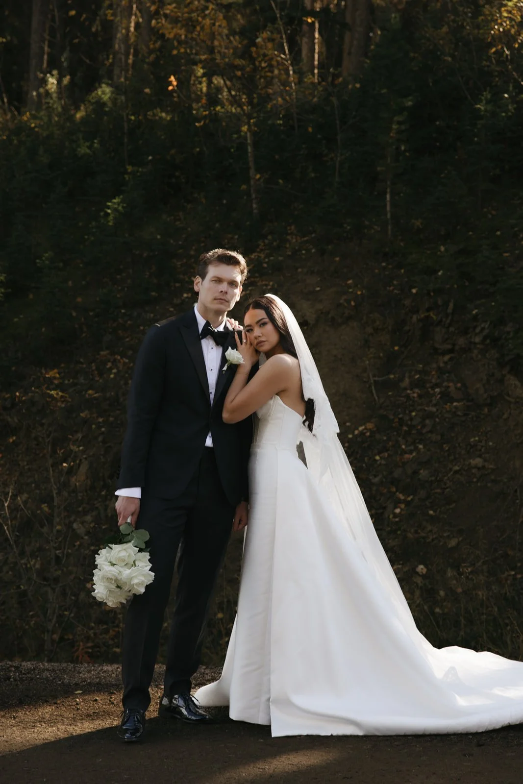 A bride and groom standing close together outdoors, with the bride in a long white wedding dress and veil, and the groom in a tuxedo holding a bouquet of white roses, against a wooded background.