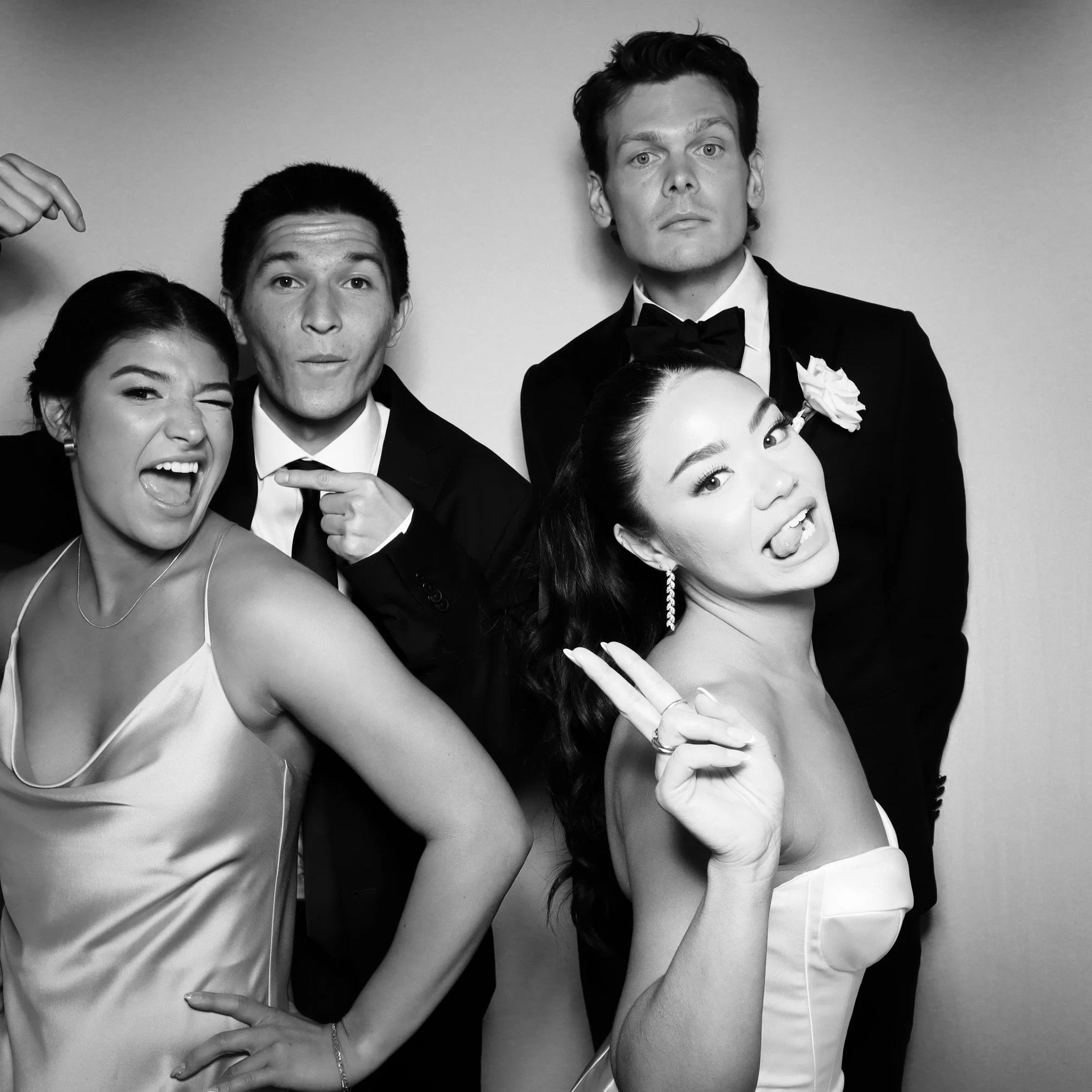 Group of four young adults in formal attire posing playfully, with two women and two men, some making silly faces and gestures, in black and white.