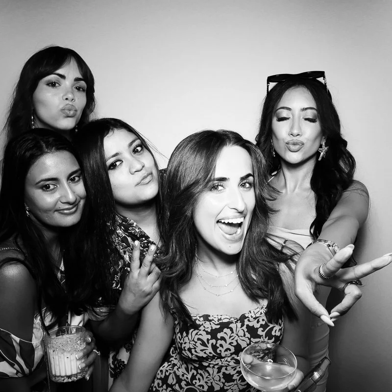 Five women at a party, taking a selfie together, posing, holding drinks, and making playful gestures, with some wearing accessories like sunglasses and jewelry.