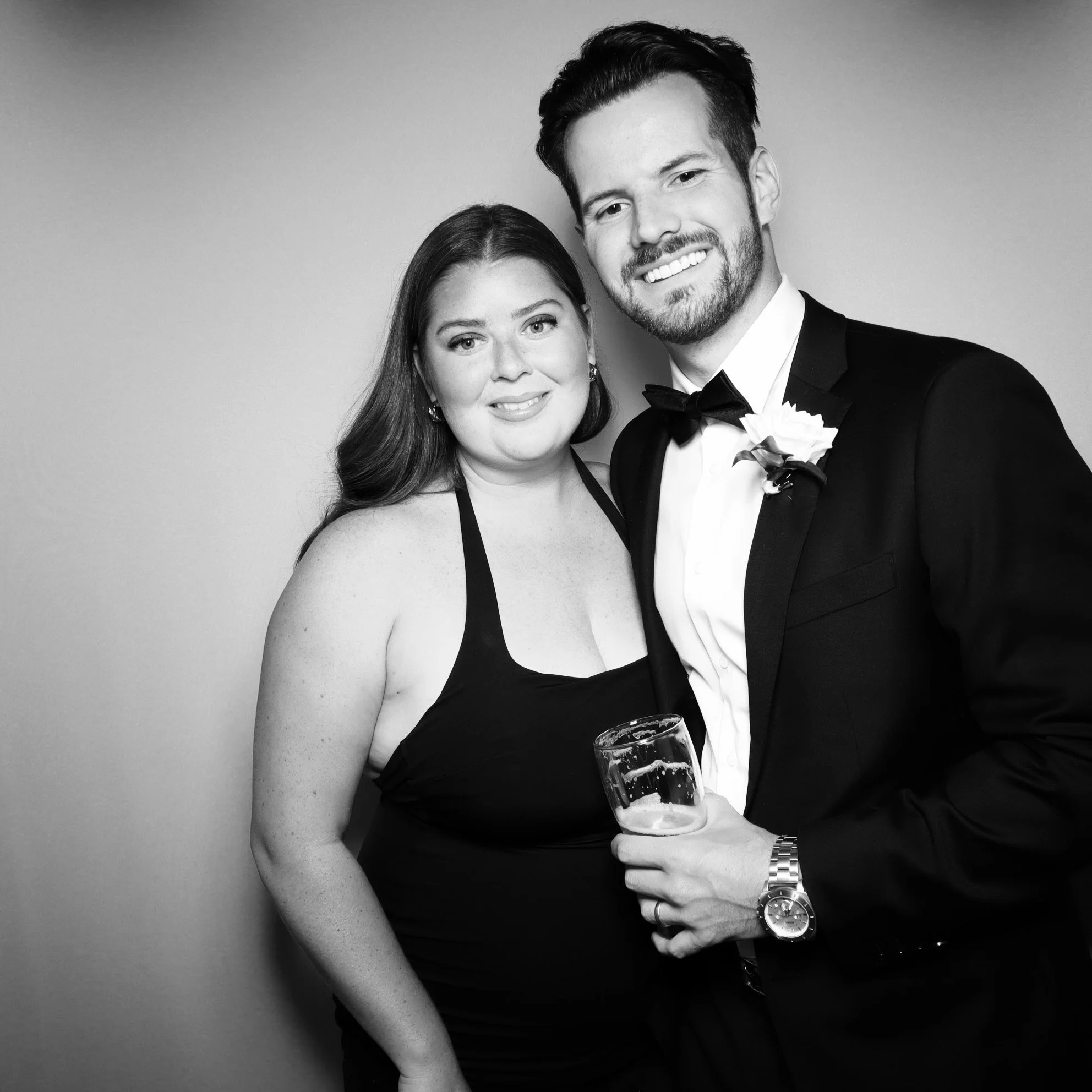 Black and white photo of a smiling couple at a formal event; woman in a halter dress and man in a tuxedo with a bow tie, holding a glass.