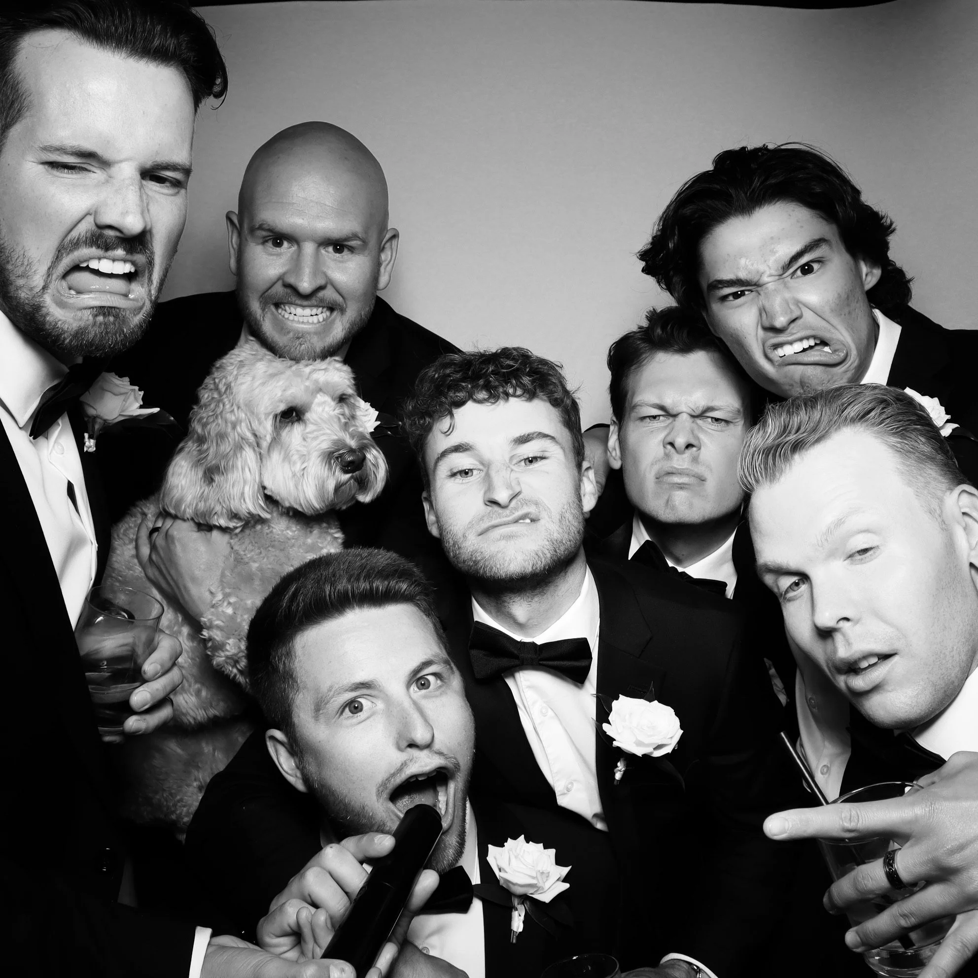 Group of men in tuxedos making various facial expressions, some angry or silly, with a dog being held by one of the men, in a black-and-white photo.