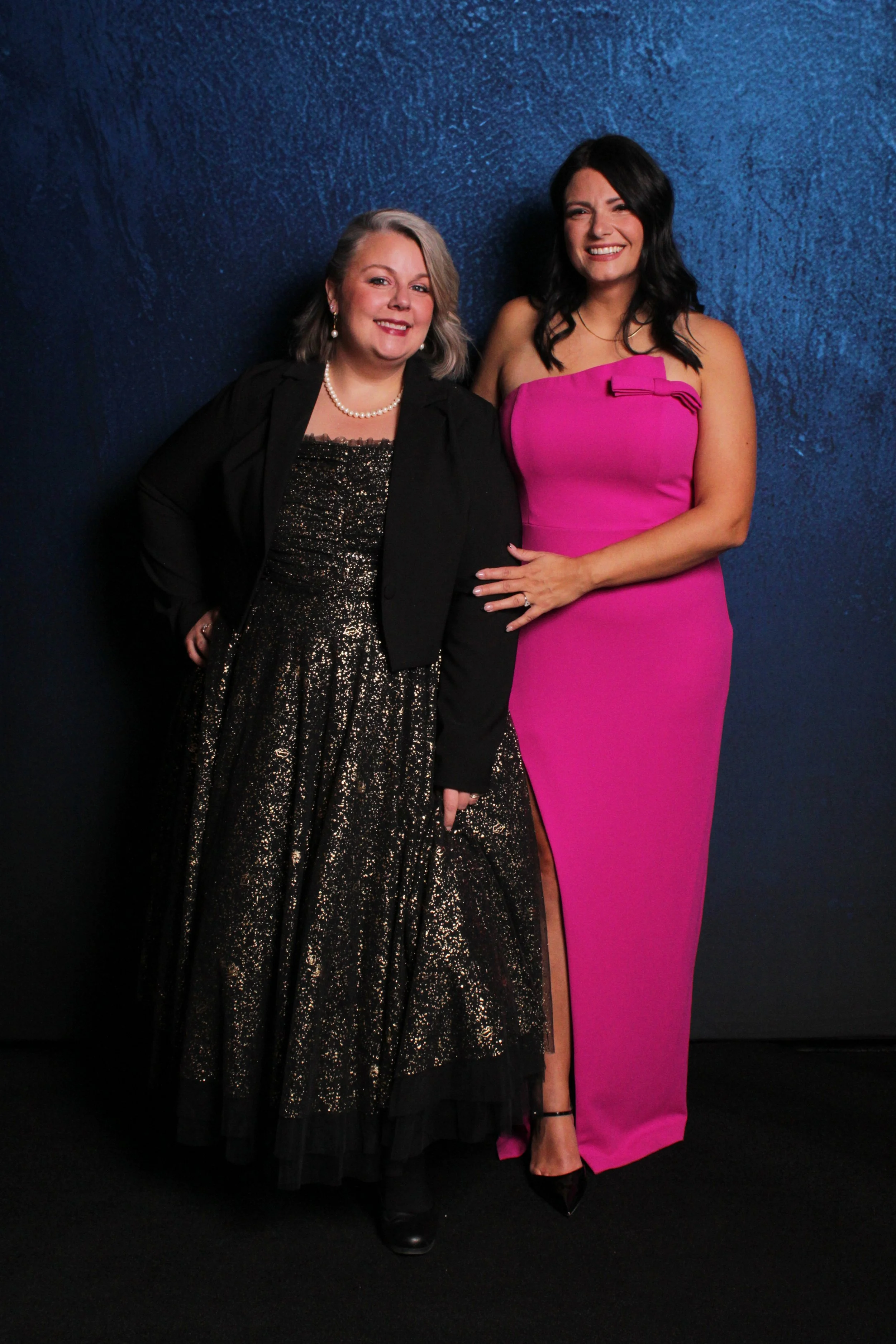 Two women in evening dresses posing against a blue textured background. The woman on the left wears a black and gold glitter gown with a matching black blazer and pearl necklace. The woman on the right wears a bright pink strapless dress with a slit,
