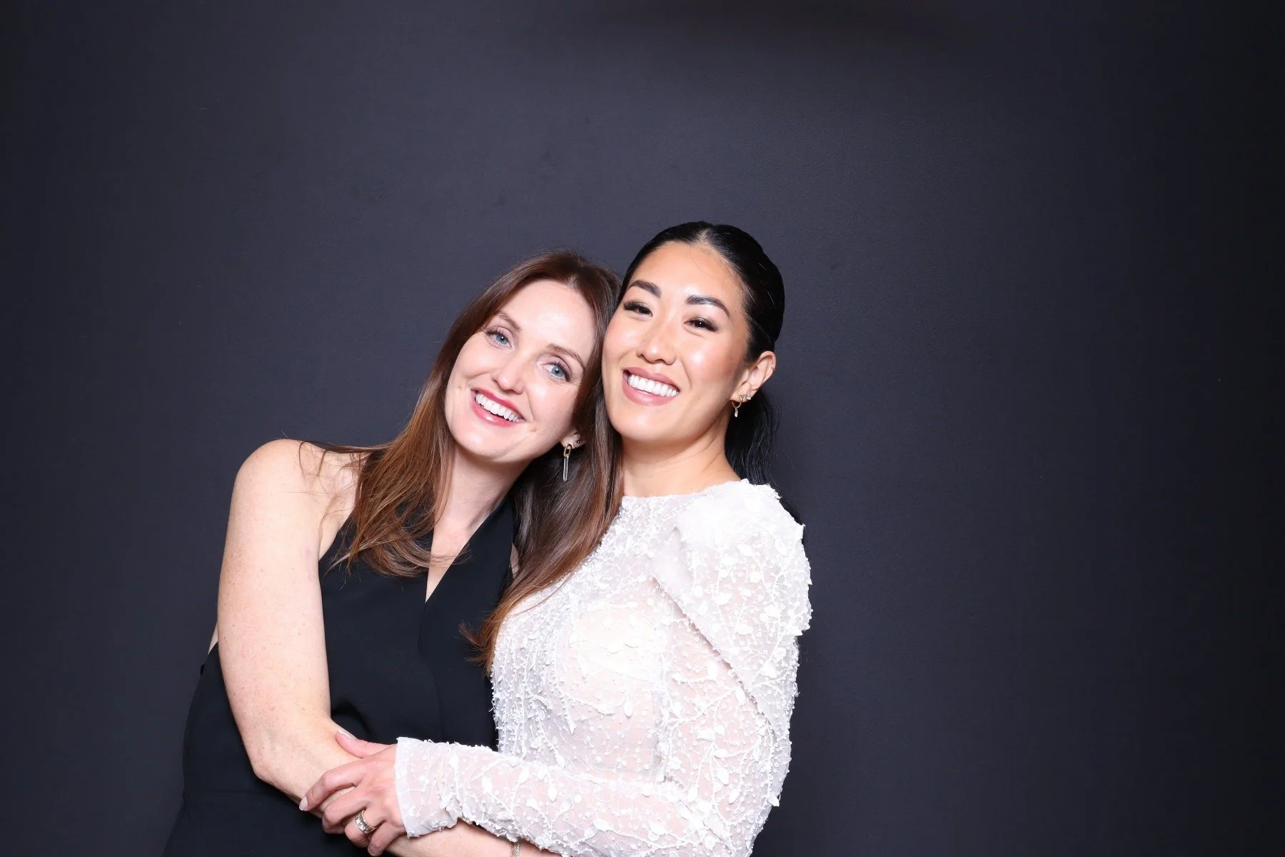 Two women posing together against a dark background. The woman on the left has long brown hair and is wearing a black top, smiling with her head slightly tilted. The woman on the right has black hair styled back and is wearing a white lace dress, smi