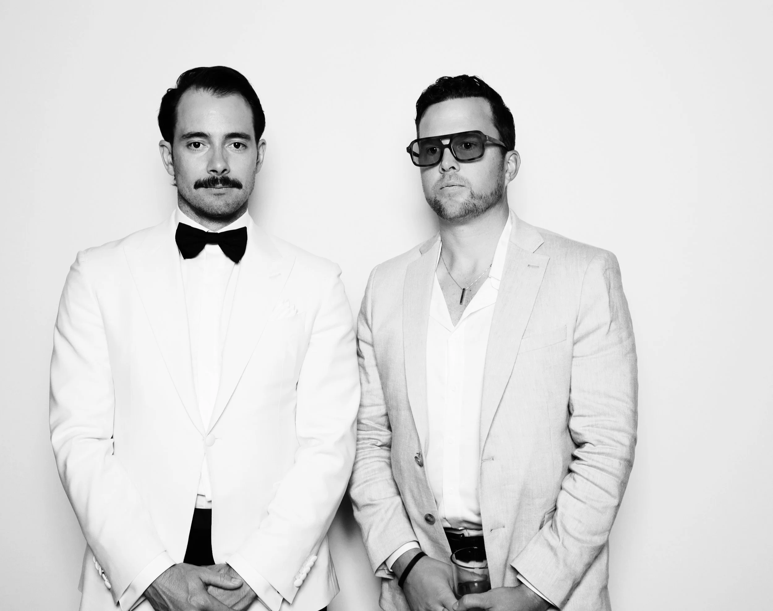 Two men dressed in formal attire standing against a plain background, one in a white tuxedo with a bow tie and the other in a light-colored blazer with glasses and holding a drink.