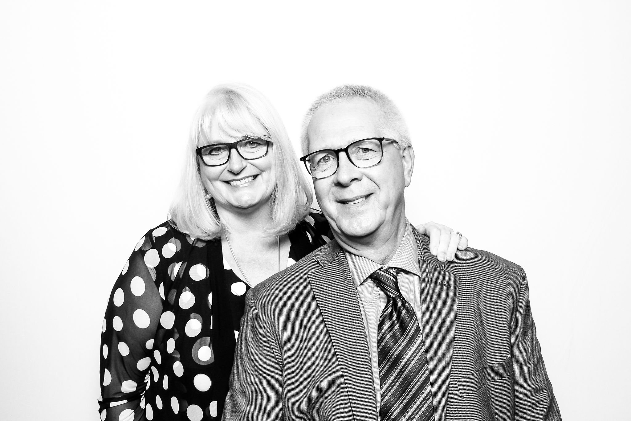 Black and white portrait of a smiling middle-aged woman with shoulder-length blonde hair, glasses, wearing a polka dot blouse, and a middle-aged man with short light hair, glasses, wearing a suit and striped tie, standing together against a plain whi
