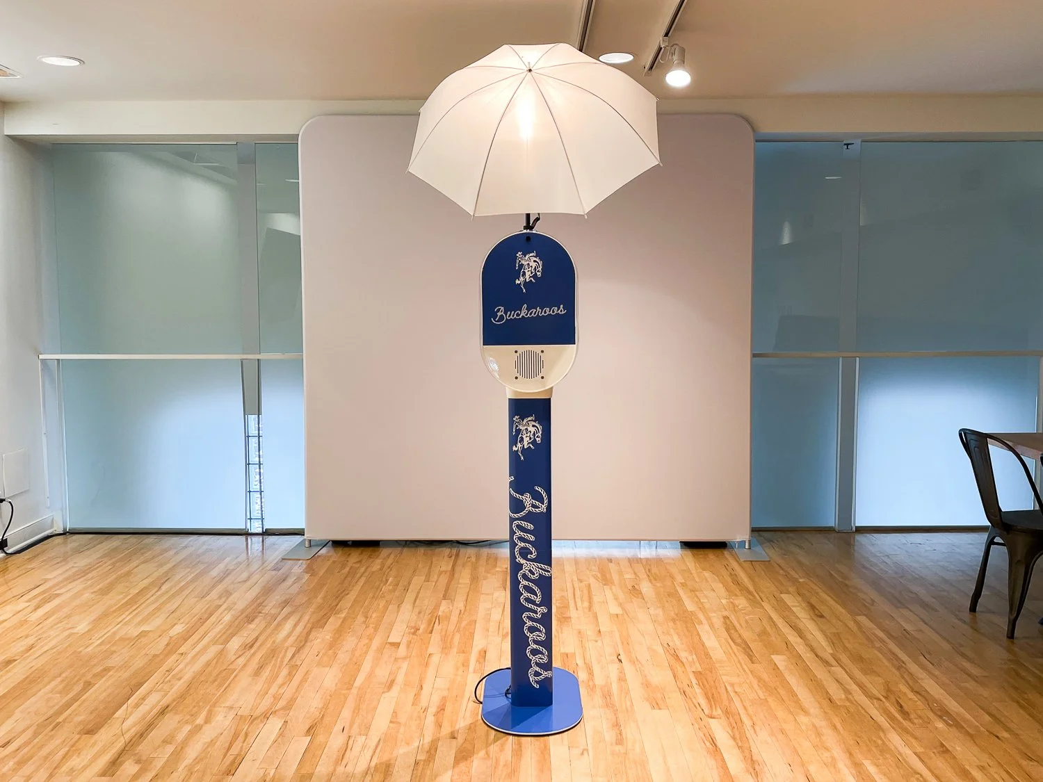 A decorative floor lamp with an open white umbrella on top, featuring a blue and white surfboard-style stand with the words "Buckaroos" and illustrations of bucking broncos, placed on a wooden floor indoors.