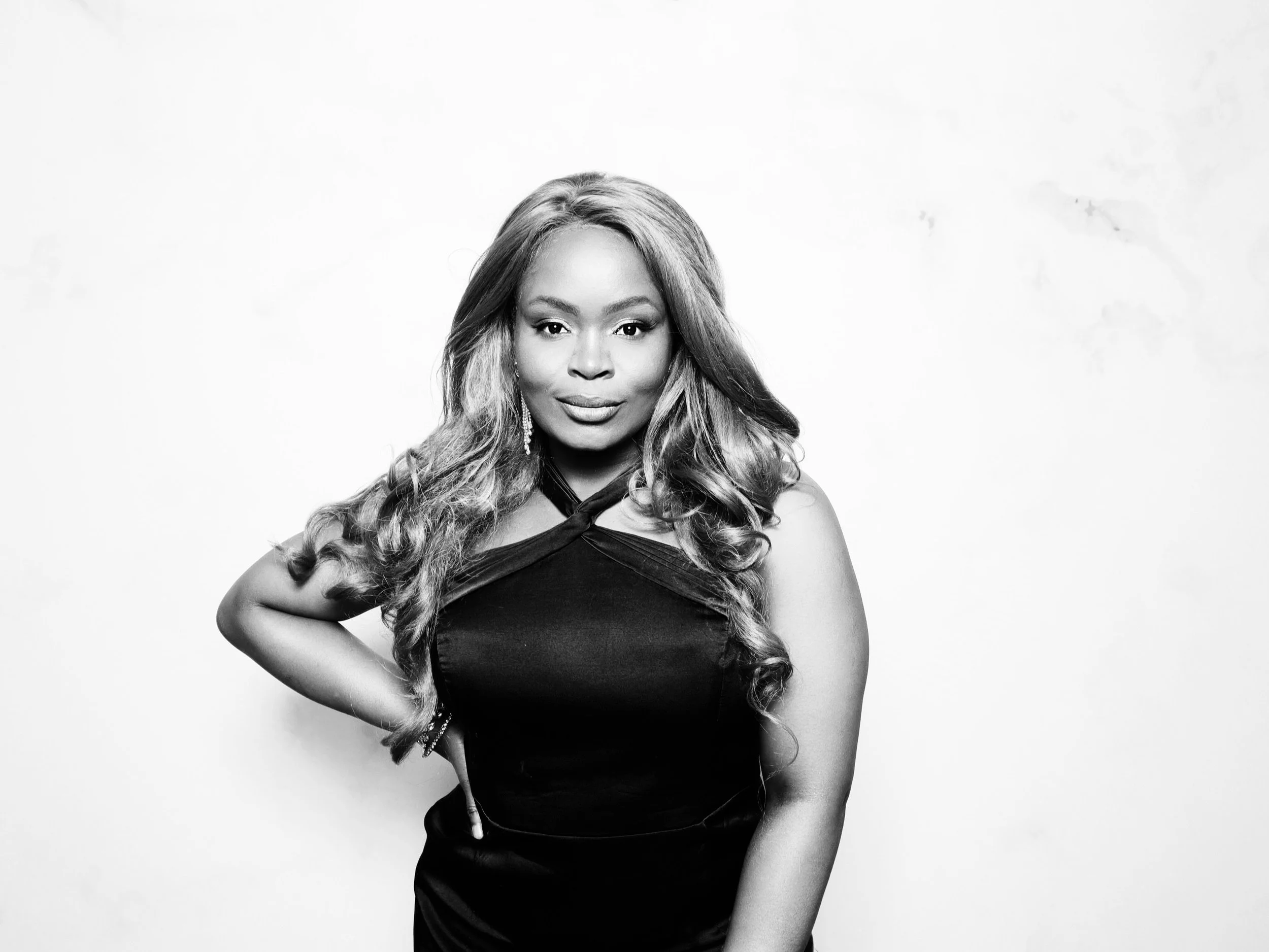 Black and white portrait of a woman with long curly hair, wearing a sleeveless black dress and earrings, standing against a plain white background.