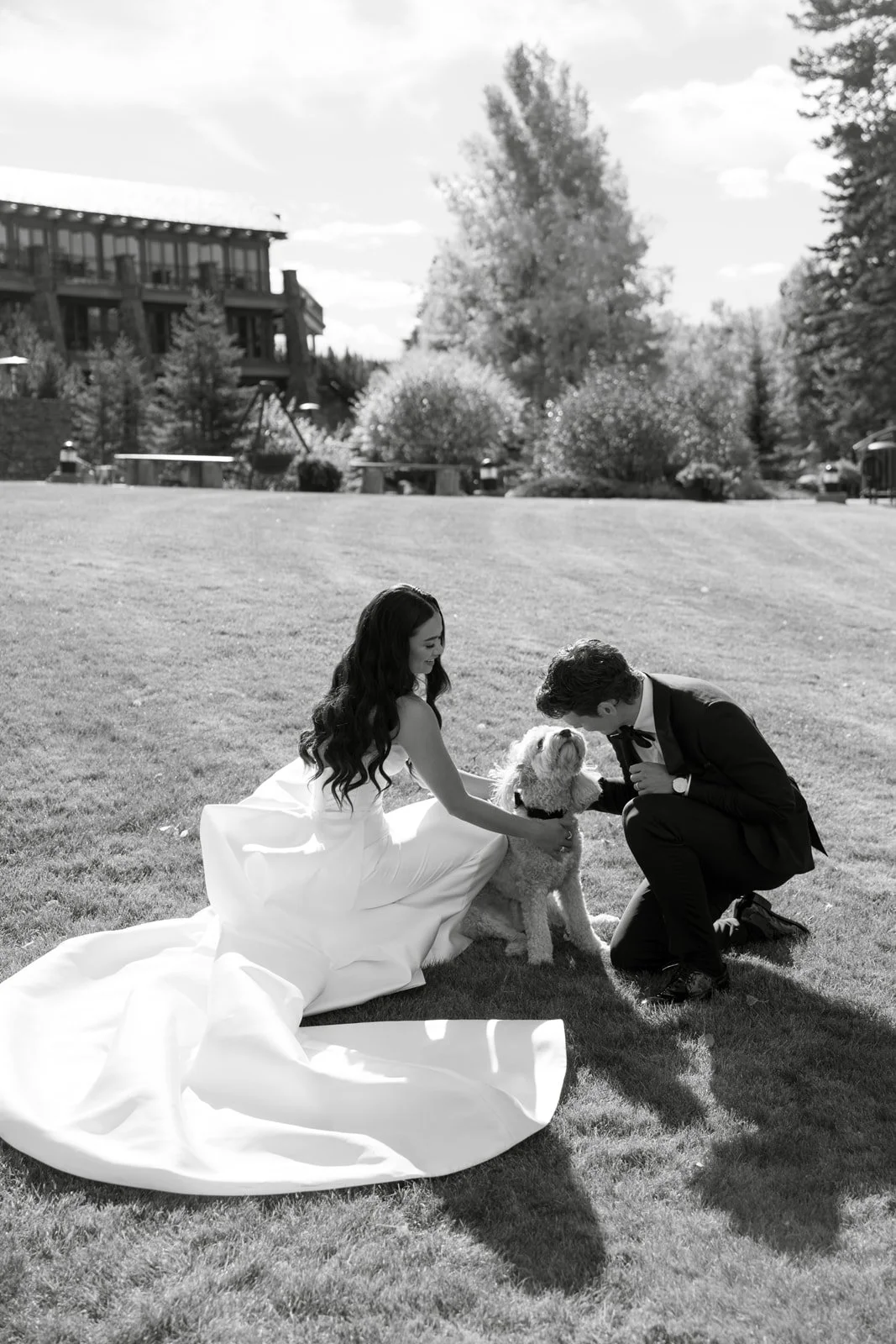 A woman in a wedding dress and a man in a suit are outdoors on a grassy area, kneeling and petting a dog, with a house and trees in the background.