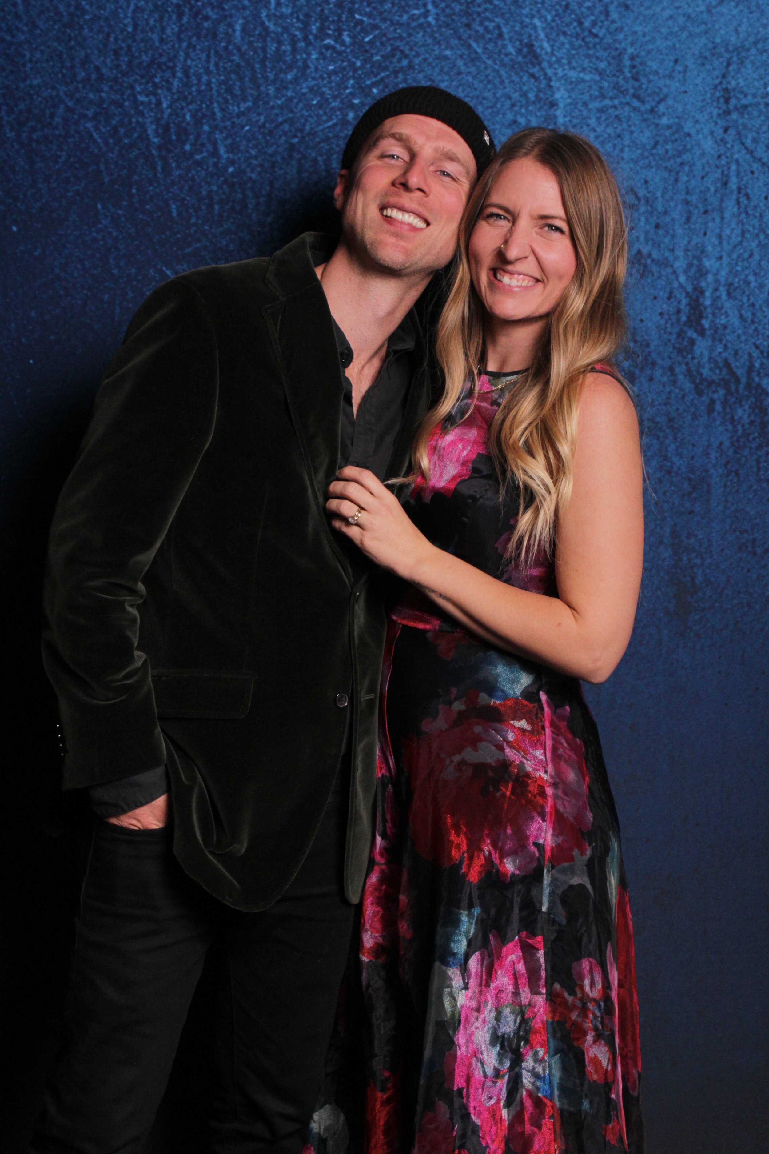 A smiling couple standing against a blue textured background, with the man in a black blazer and beanie, and the woman in a colorful floral dress.