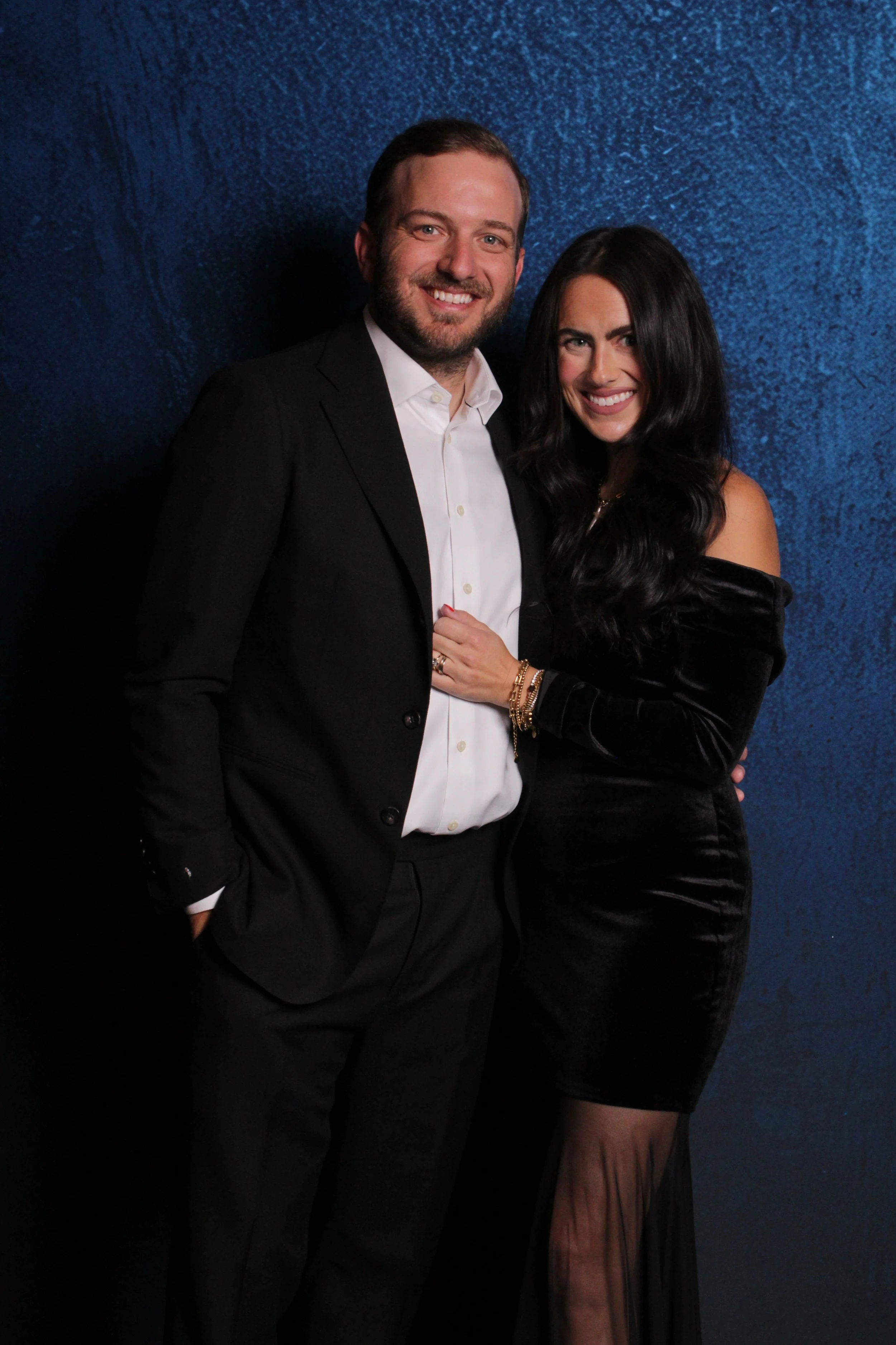 A man and woman dressed in formal attire, smiling and posing together against a textured blue background.