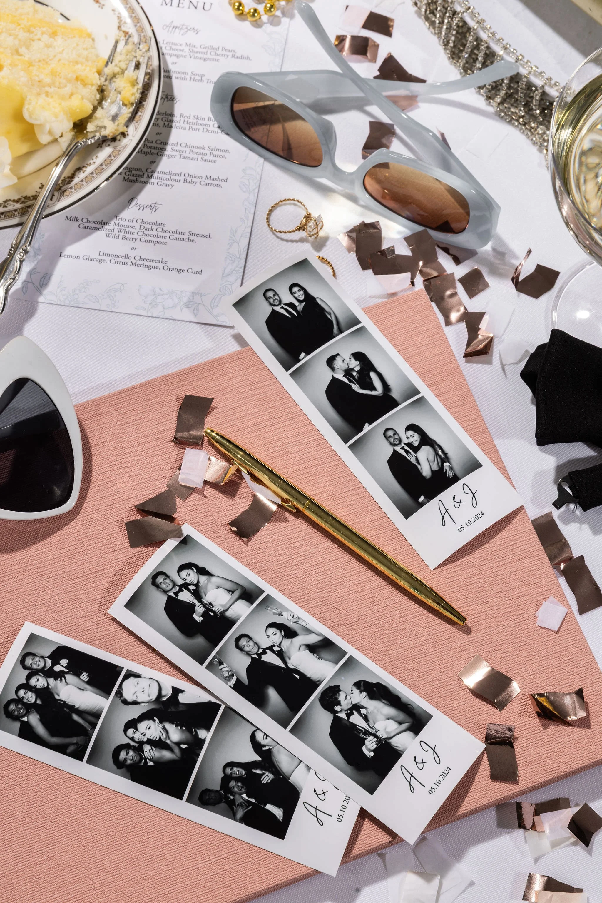 A table at a wedding reception with black-and-white photo booth strips, a gold pen, sunglasses, a bracelet, and scattered rose gold and white confetti. A wedding menu and a champagne glass are also visible.