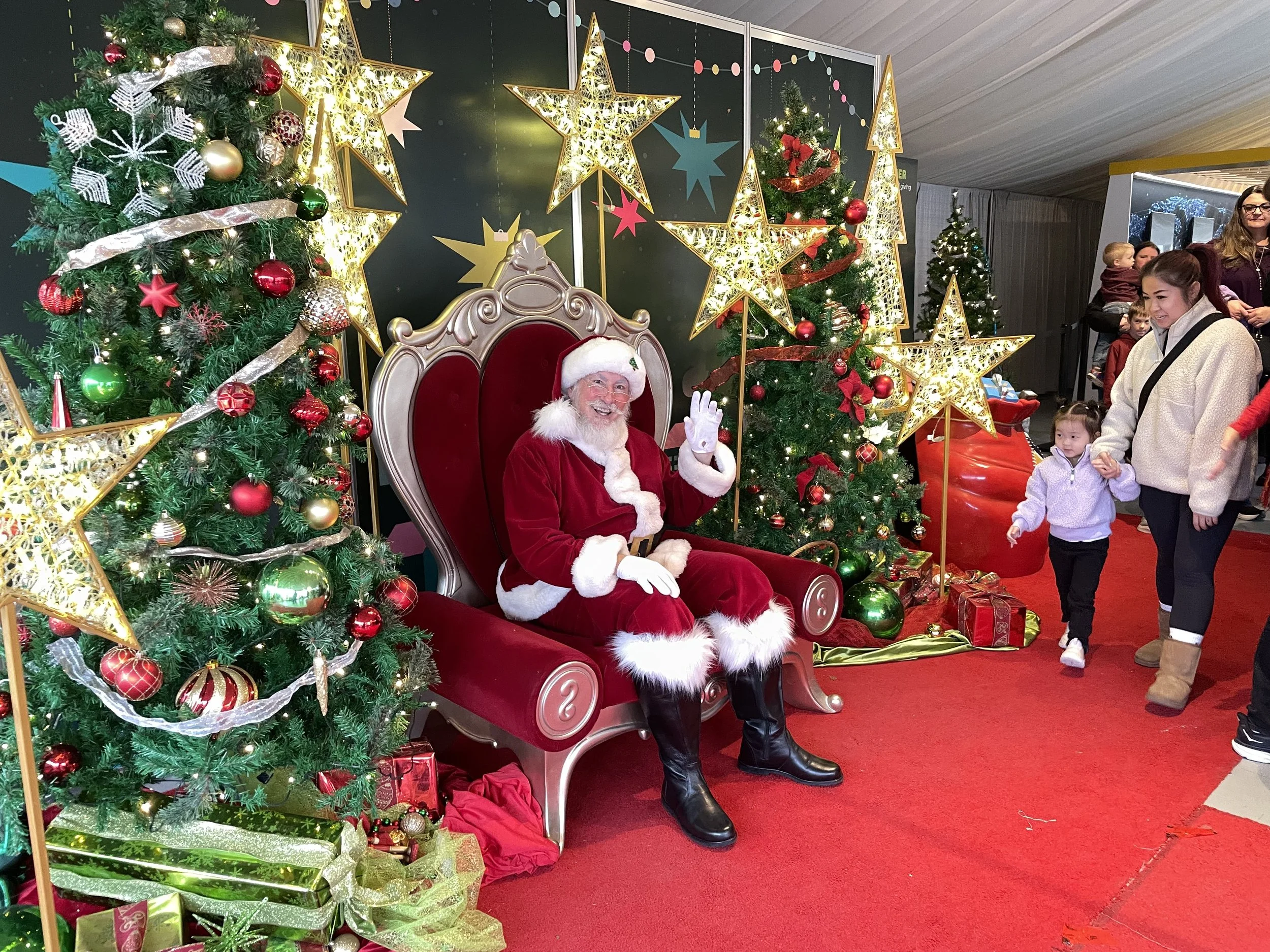 Santa Claus sitting on a throne, waving, at a Christmas event with decorated trees and star ornaments, with children and adults nearby.