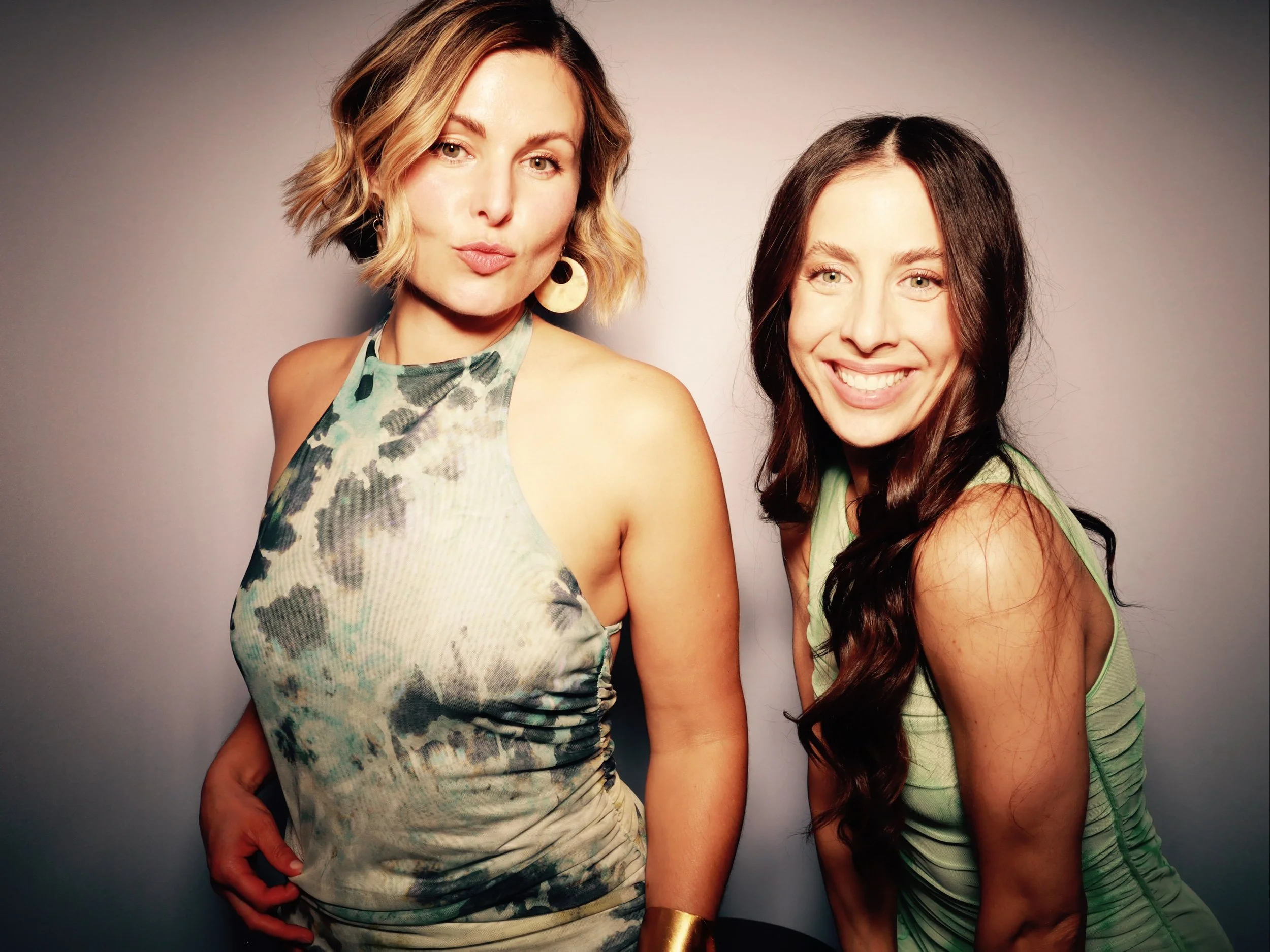 Two women smiling and posing for a photo against a plain background. The woman on the left has blonde hair with waves, wears a sleeveless patterned top, and has gold hoop earrings and a gold bracelet. The woman on the right has long dark hair, wears 