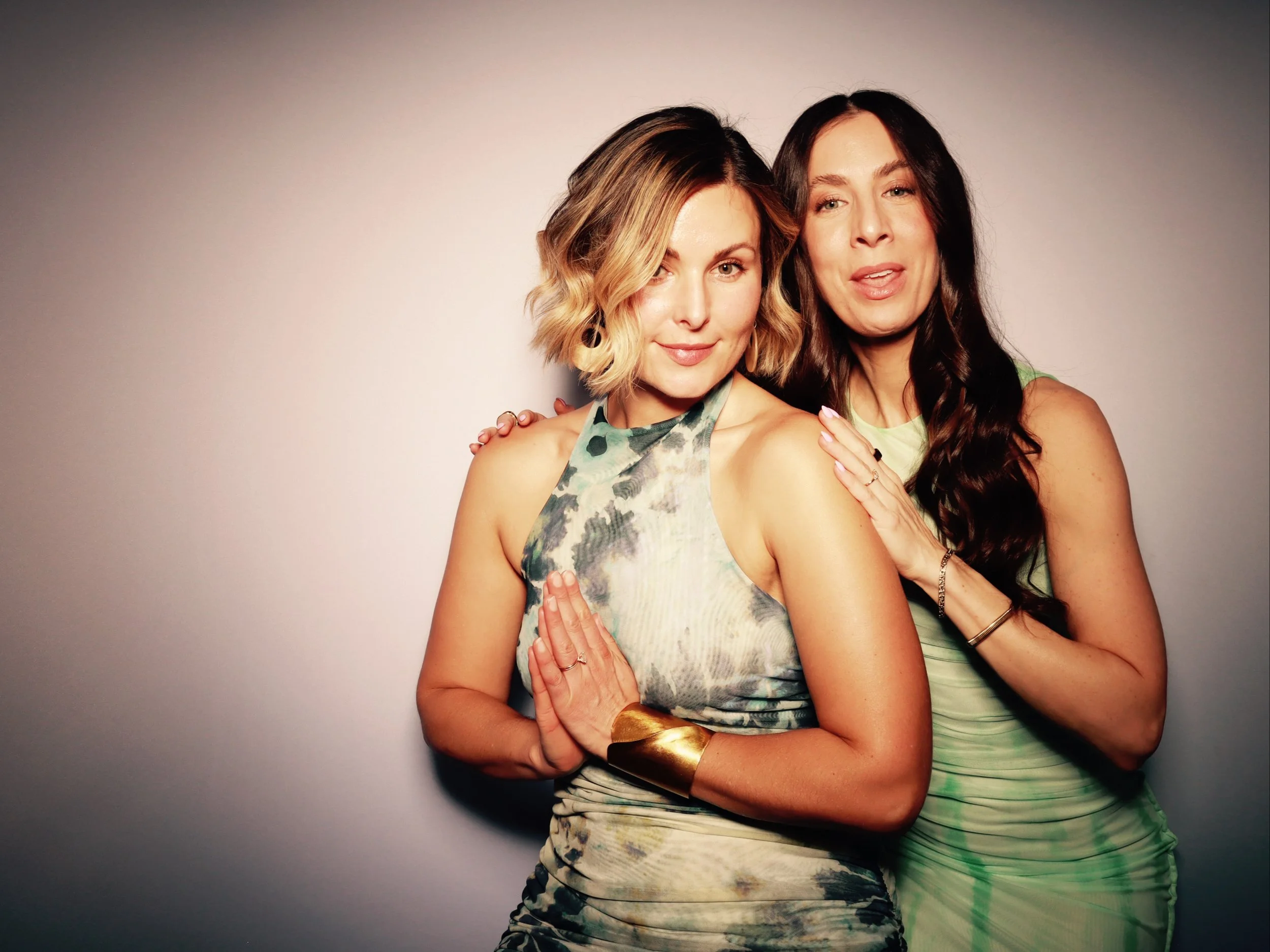 Two women posing together, one with short wavy blonde hair and a sleeveless patterned dress, the other with long straight dark hair and a sleeveless green dress, standing closely with hands on each other, against a neutral background.