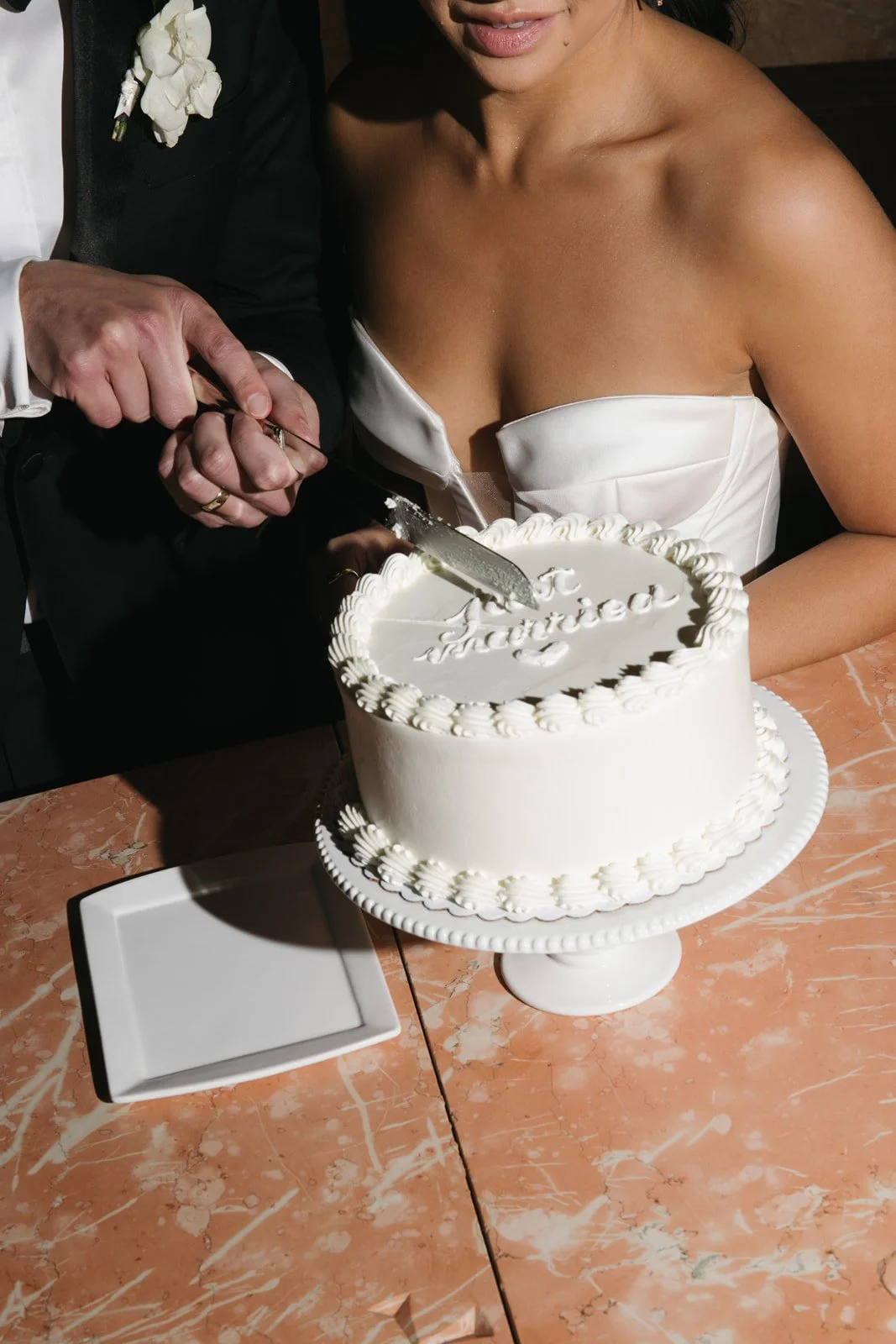 A bride in a white strapless dress is cutting a wedding cake with a man dressed in a black suit and white shirt. The cake is white with decorative icing and reading 'Just Married' on top.