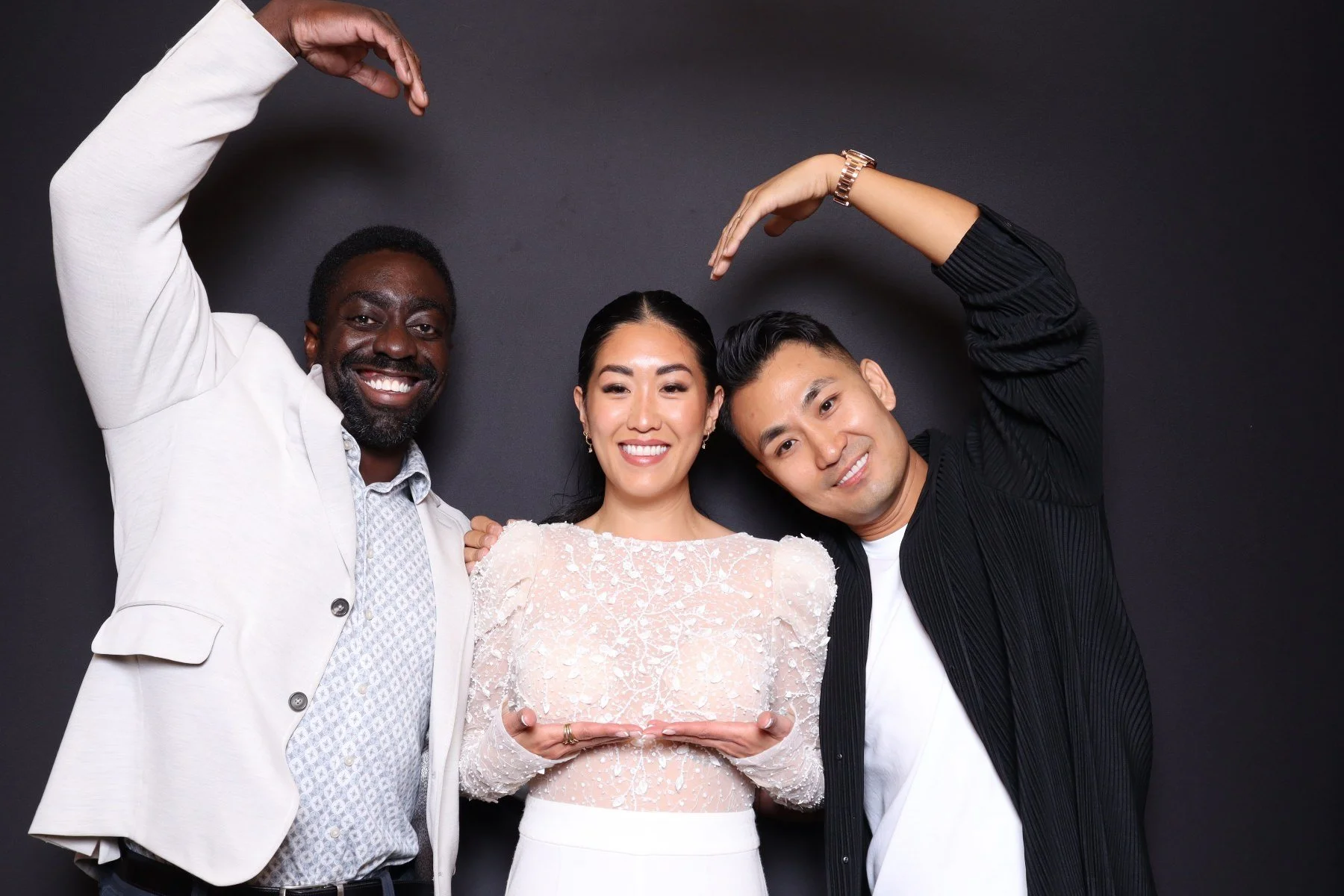 Three people smiling and forming a heart with their arms against a dark background. The woman in the middle holds a clear plaque.